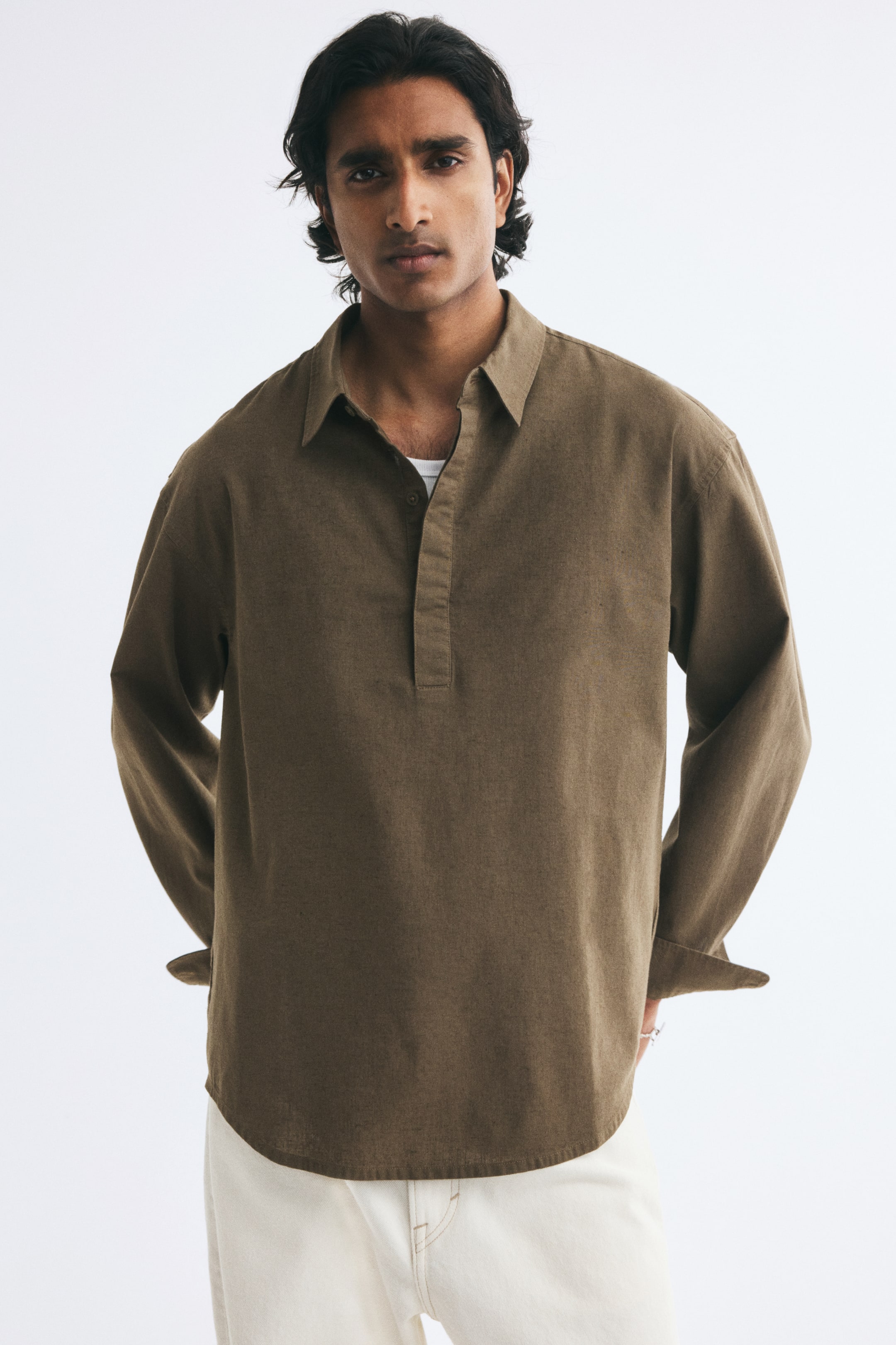 View larger image: The model is wearing a brown linen-blend popover shirt with a relaxed fit, a classic collar, and a partially open placket. The long-sleeved shirt features dropped shoulders and a rounded hem, styled over a white undershirt and light-coloured trousers.