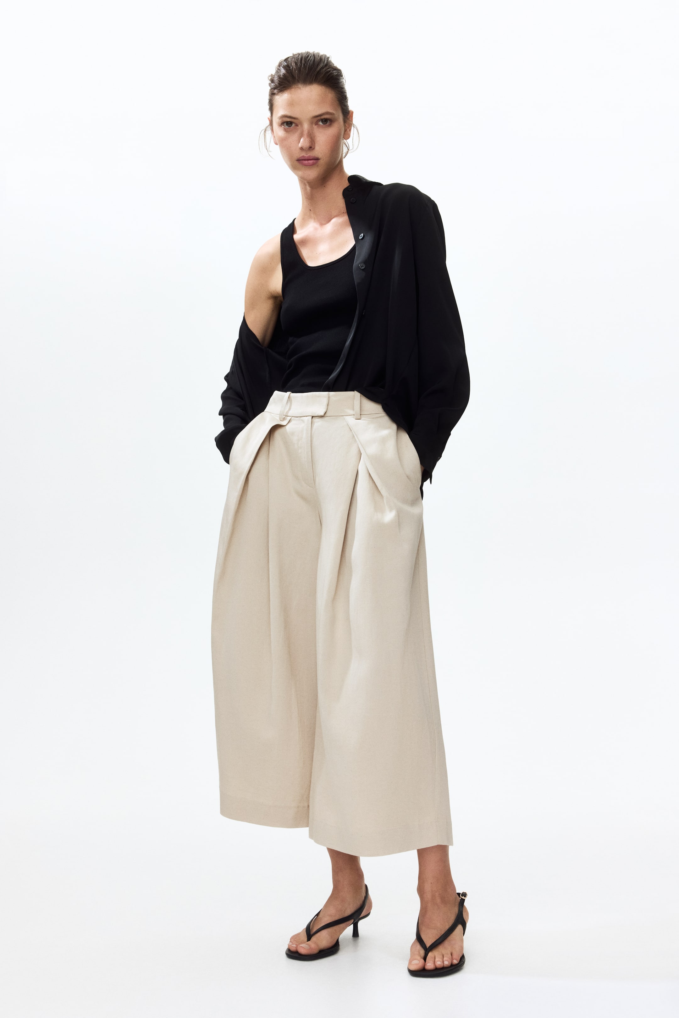 View larger image: Beige wide-leg trousers feature prominent pleats starting from the high-waisted band, with hands casually tucked into the side pockets. These trousers offer a relaxed, cropped fit, finishing just above the ankles.