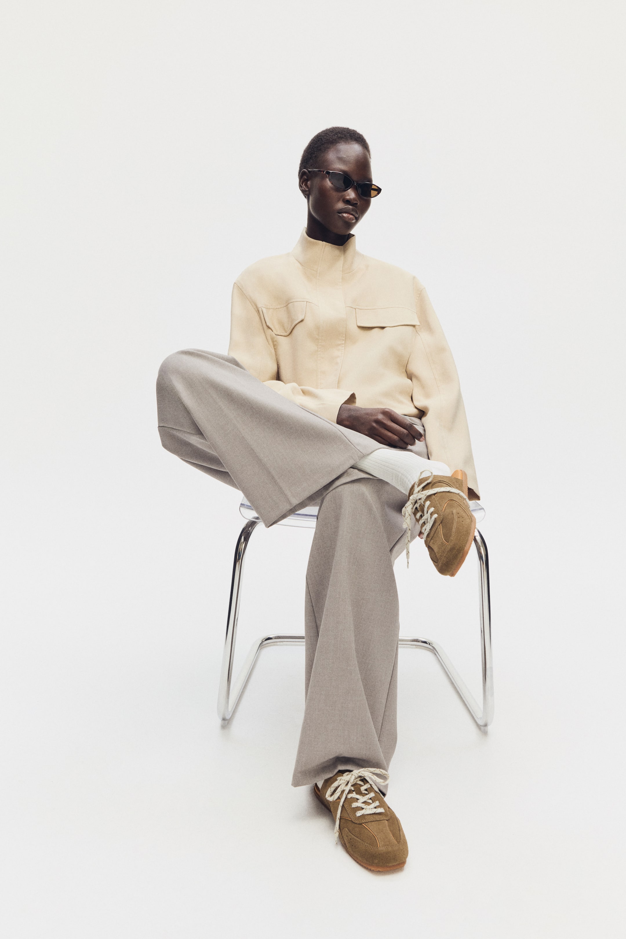 Ampliar la imagen: Las mulas estilo "trainer" en gamuza marrón suave con sutiles detalles de cuero están siendo usadas por la modelo. Presentan cordones blancos en la parte delantera y una lengüeta acolchada, con la modelo mostrándolos sobre calcetines blancos mientras cruza una pierna.