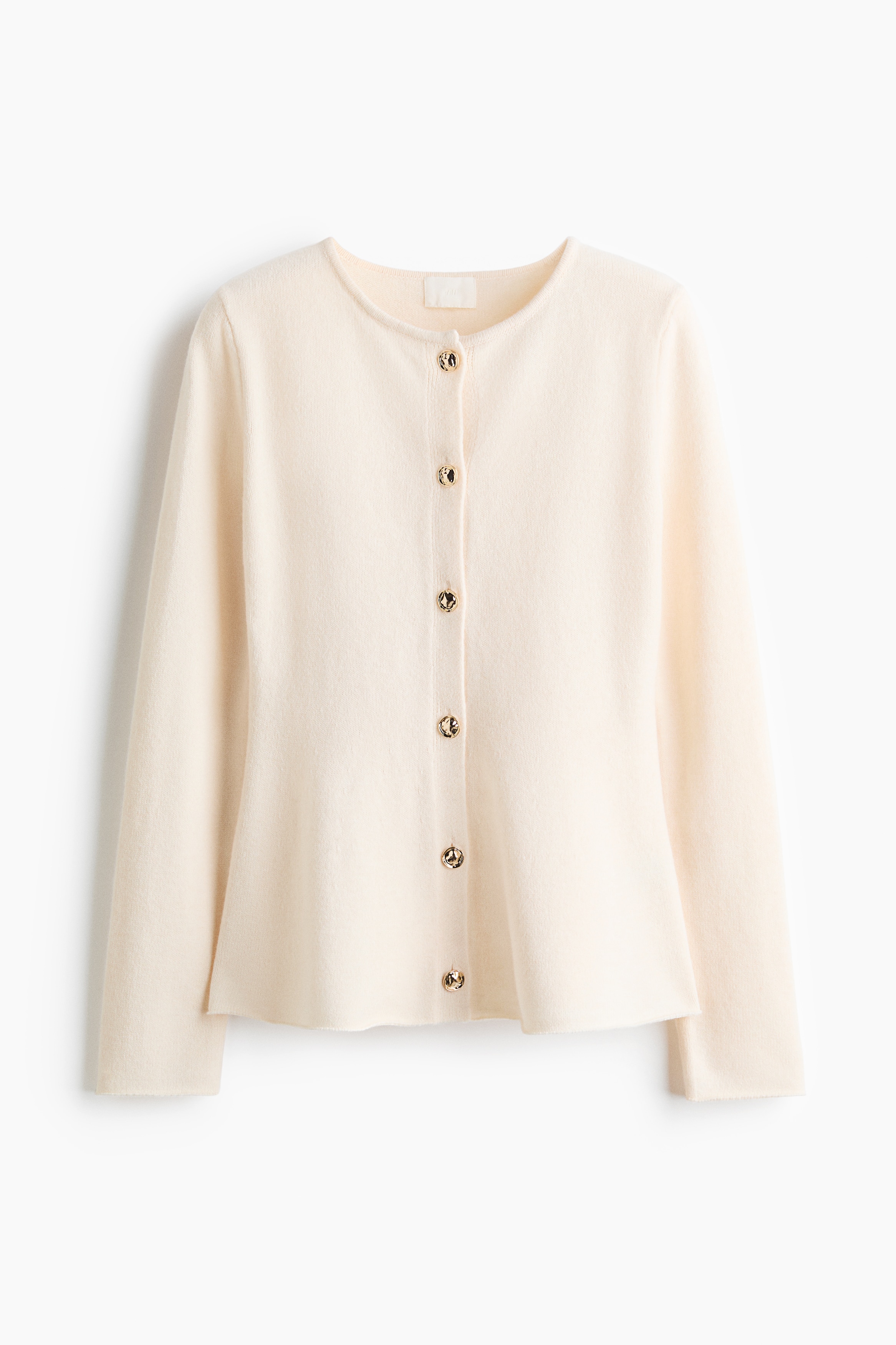 A cream-coloured knit cardigan featuring a round neckline, front button fastenings, long sleeves, and a flared peplum hem is being worn by the model. The cardigan is styled with black leather trousers and black pointed-toe boots.