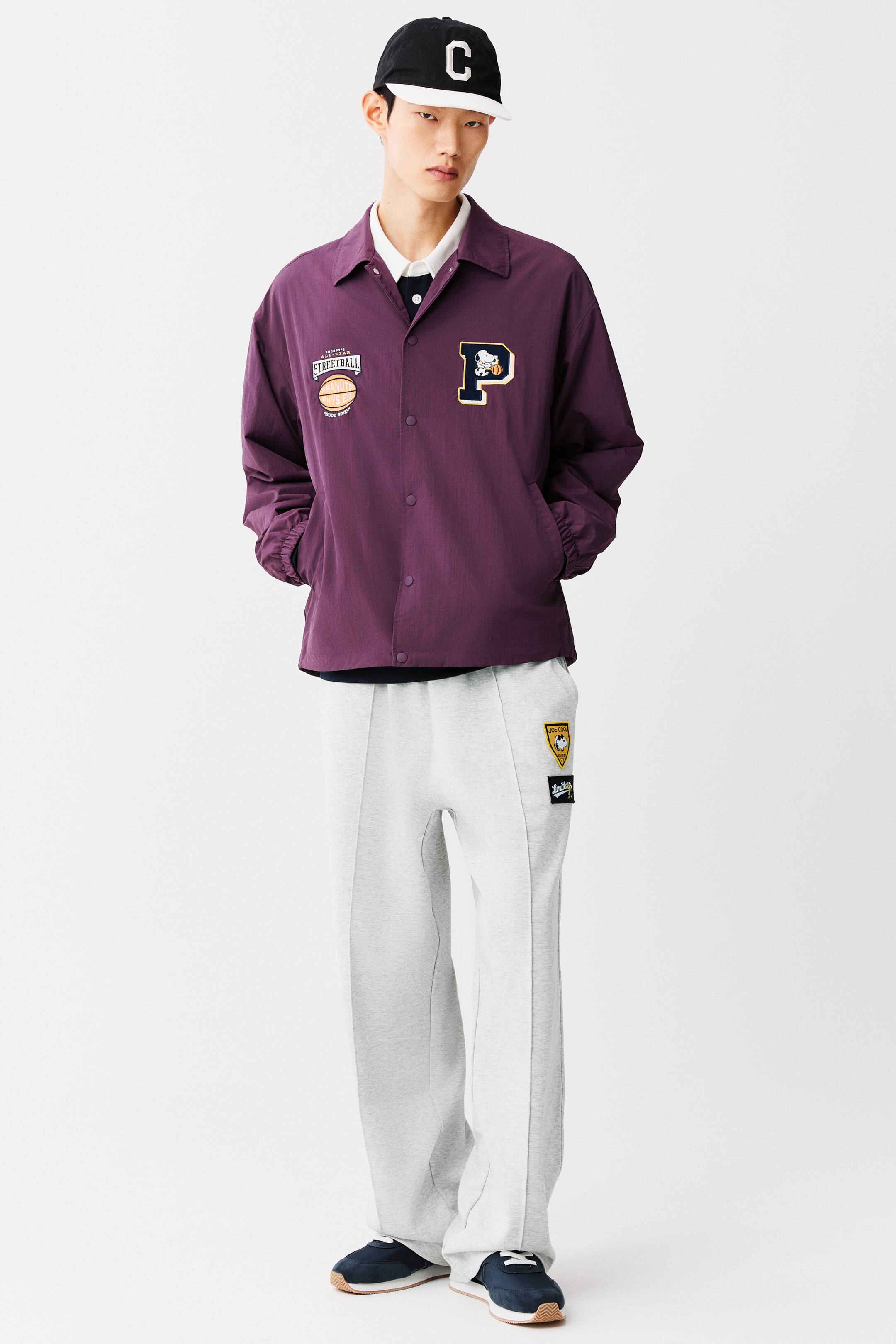 View larger image: A purple coach jacket featuring a snap-button closure, a classic collar, and graphic patches on the chest is worn by the model, who has both hands resting in the side pockets. The jacket is layered over a white collared shirt and paired with light grey sweatpants and a black baseball cap.
