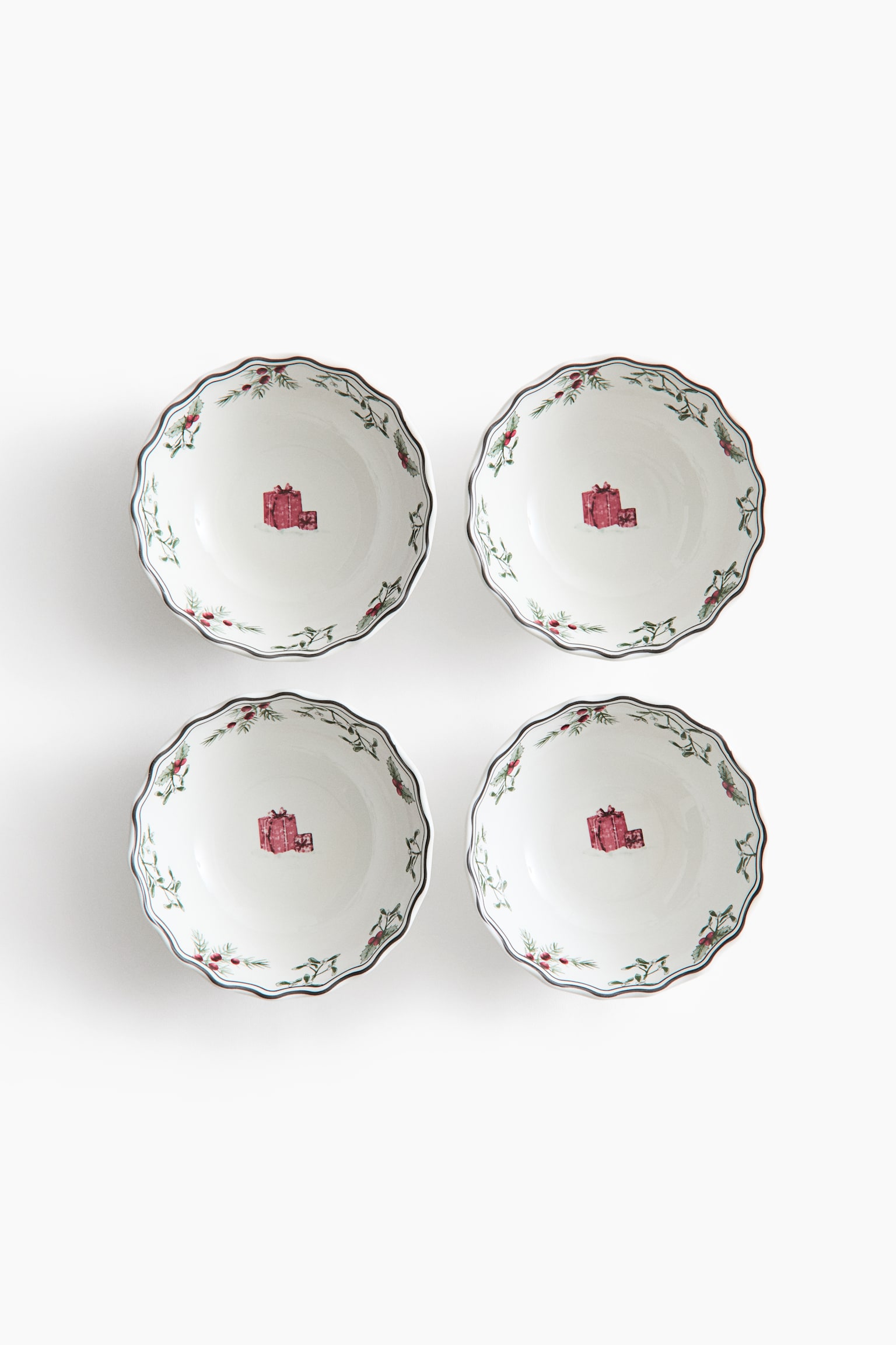 A set of four white ceramic bowls is displayed from a top-down perspective, featuring scalloped rims with a dark border. Each bowl showcases a central illustration of red wrapped gift boxes, complemented by a delicate border of green evergreen branches and red berries along the inner rim. - 1