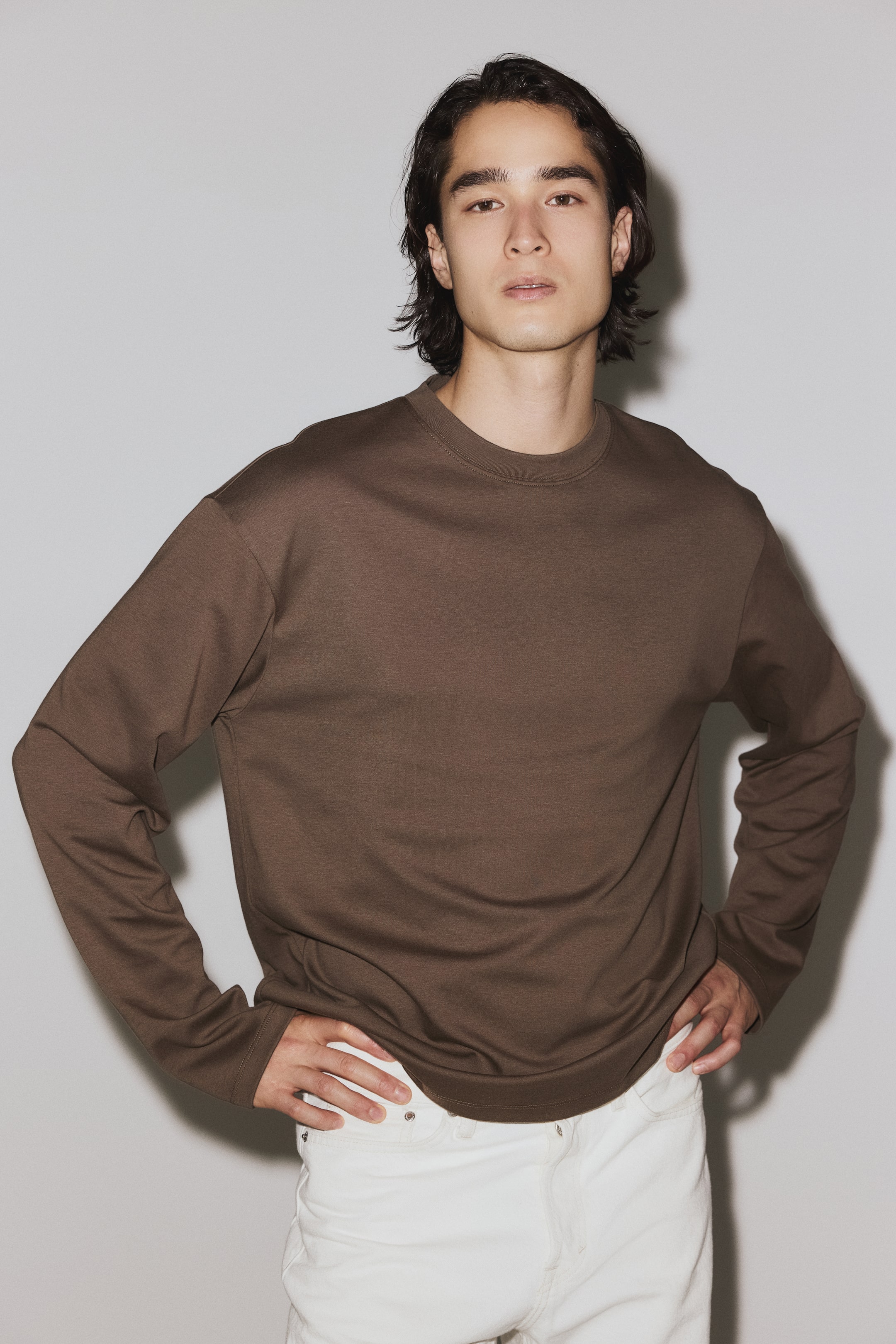 Visualizza immagine più grande: Un top marrone a maniche lunghe con scollo girocollo è presentato su una modella vista frontalmente. È caratterizzato da un tessuto liscio, spalle scese, una vestibilità rilassata e polsini e orlo a coste.