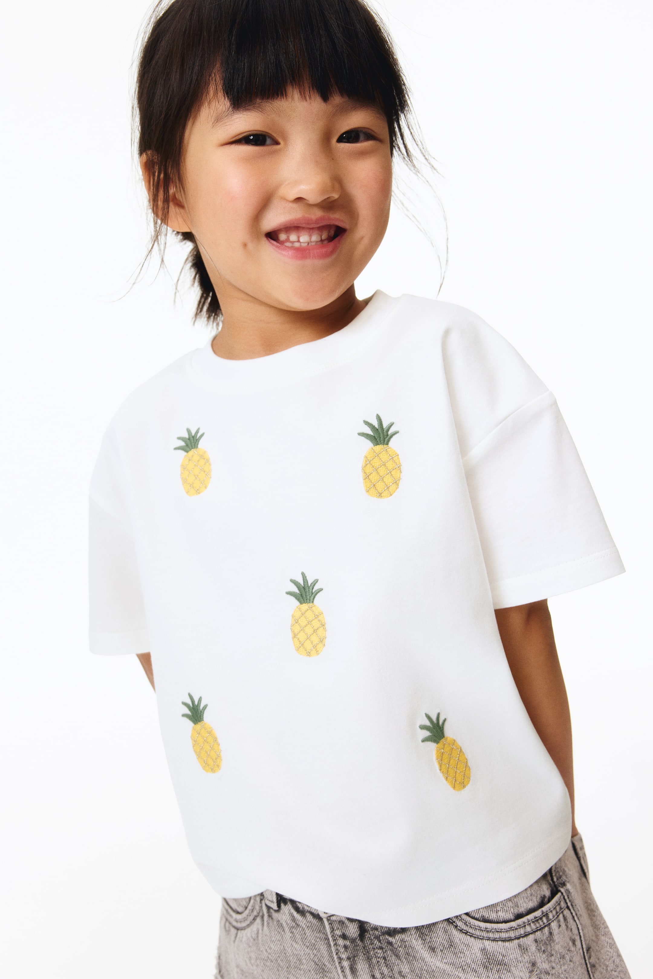 Grotere afbeelding bekijken: Een wit T-shirt toont meerdere geborduurde ananasmotieven verspreid over de voorkant, met een ronde halslijn en korte mouwen.
