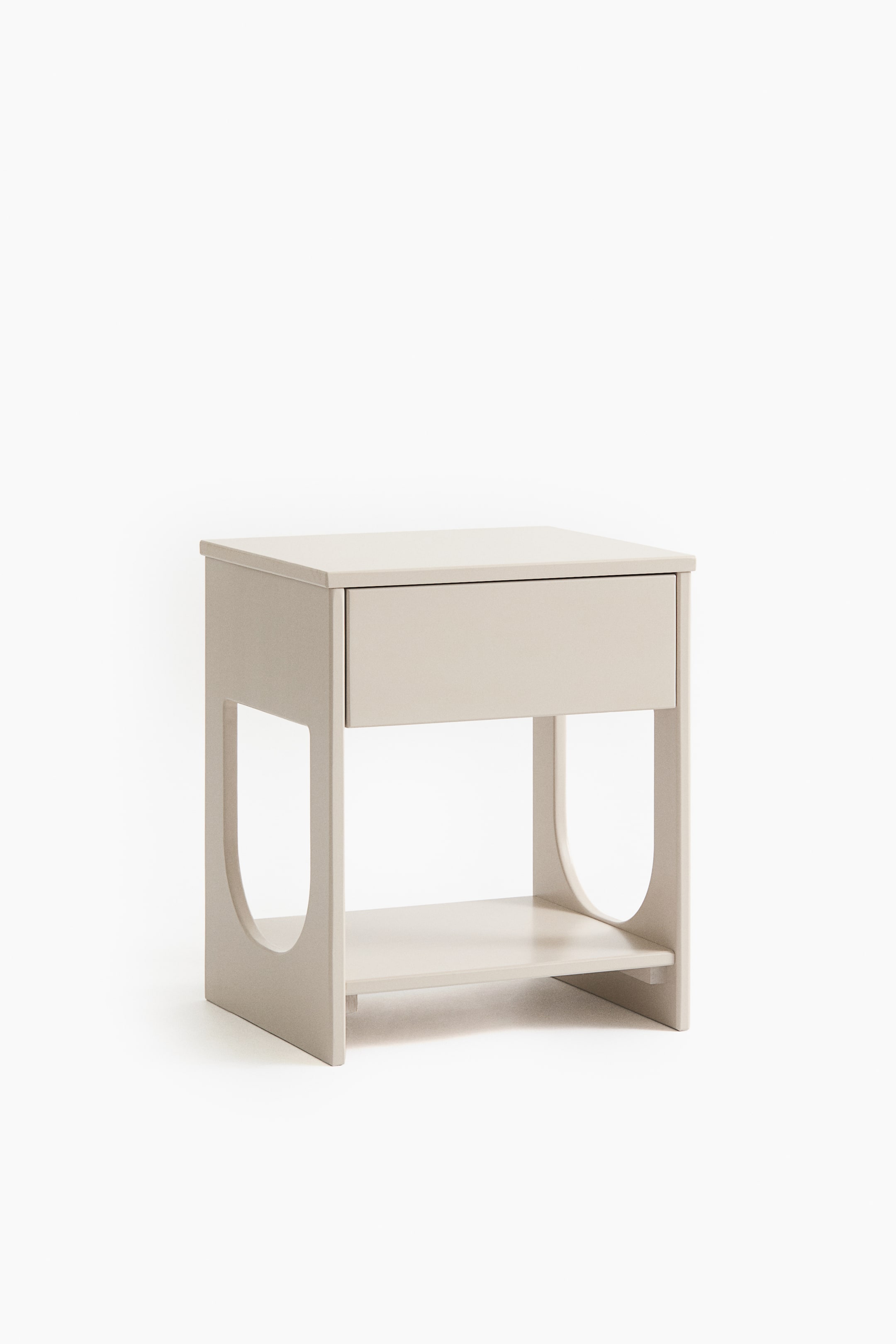 Zobraziť väčší obrázok: Svetlý béžový nočný stolík má na prednej strane jednu zásuvku, pod ňou otvorenú policu a na bočných paneloch výrezy v tvare U.