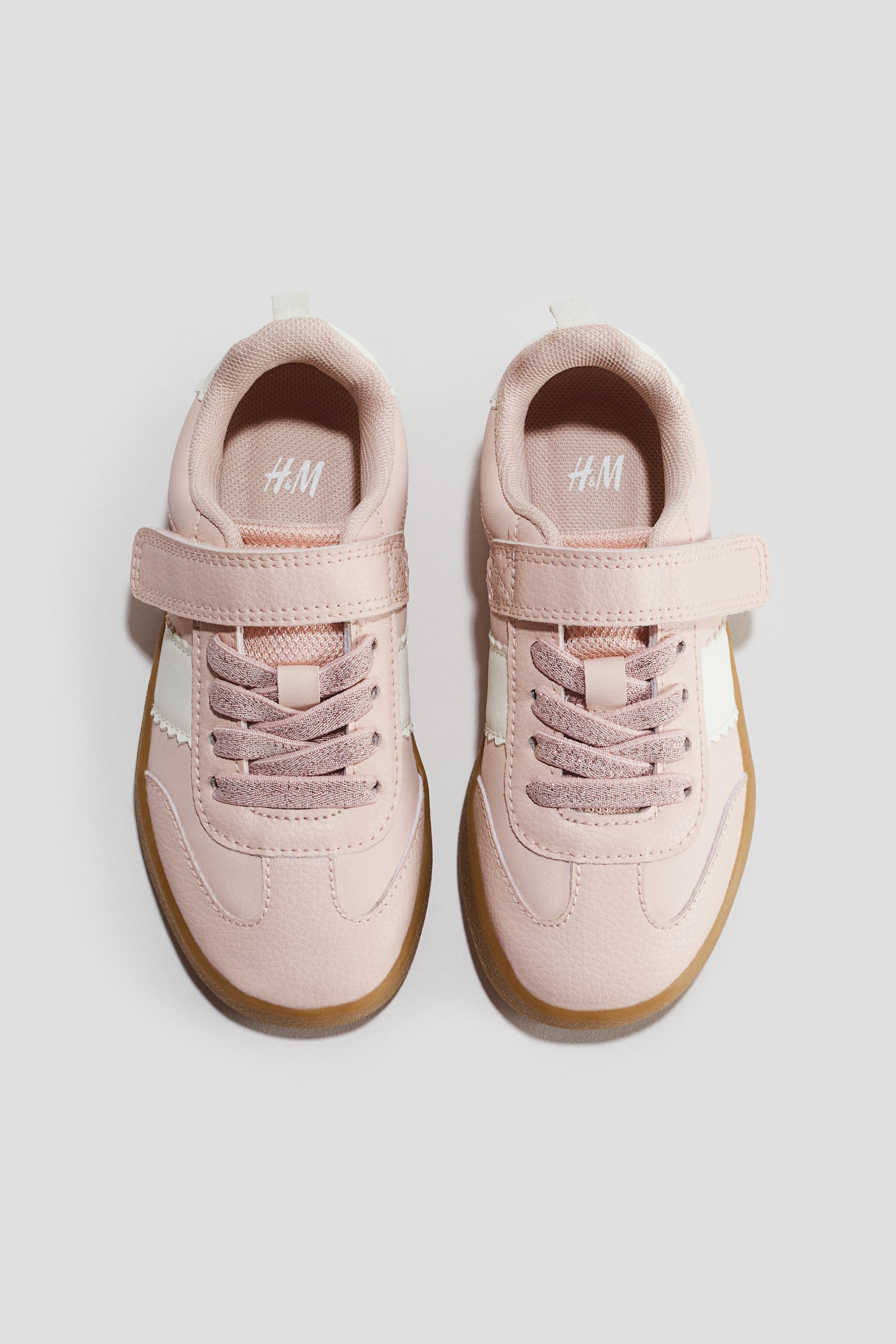 Light pink sneakers are facing forward in a top-down view, featuring a textured upper with elasticated laces and a hook-and-loop strap across the instep. The design is displaying white scalloped side panels and contrasting light brown gum soles.