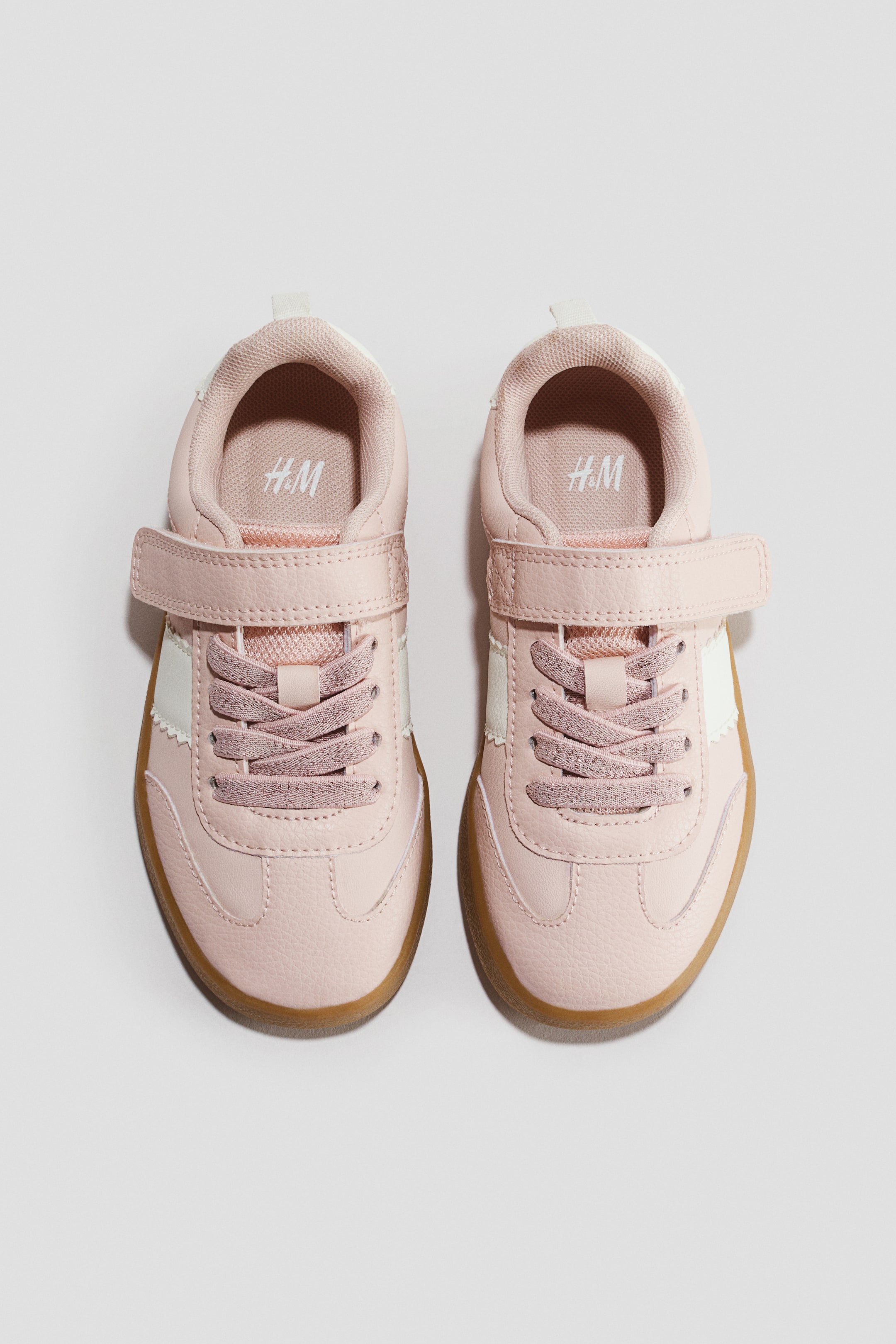 View larger image: Light pink sneakers are facing forward in a top-down view, featuring a textured upper with elasticated laces and a hook-and-loop strap across the instep. The design is displaying white scalloped side panels and contrasting light brown gum soles.