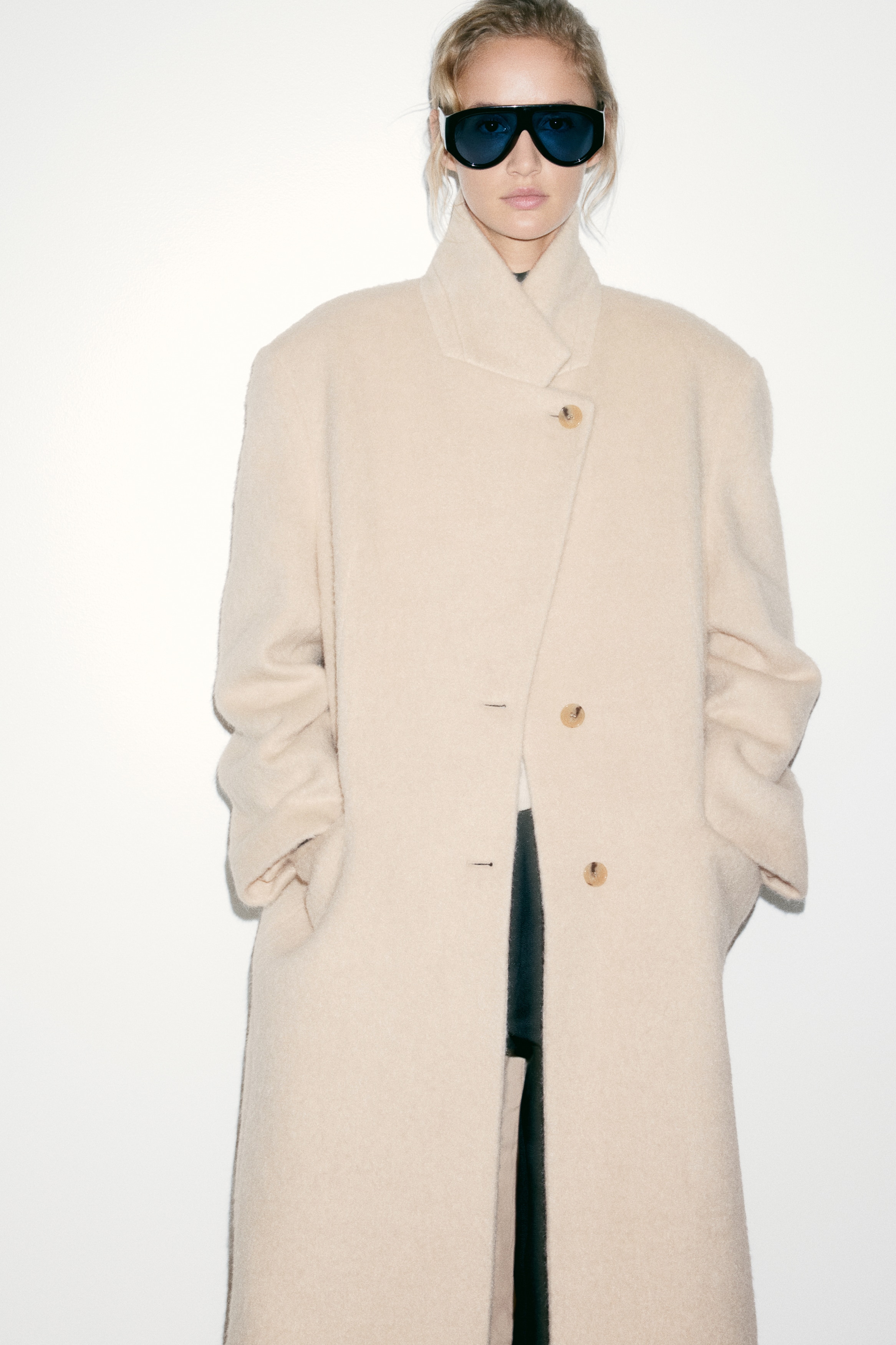 Un long manteau Ã  simple boutonnage en tissu texturÃ© beige clair est suspendu de face, mettant en valeur des revers crantÃ©s et une fermeture Ã  deux boutons. Le manteau prÃ©sente une silhouette droite avec des manches longues et deux poches passepoilÃ©es horizontales Ã  la taille.