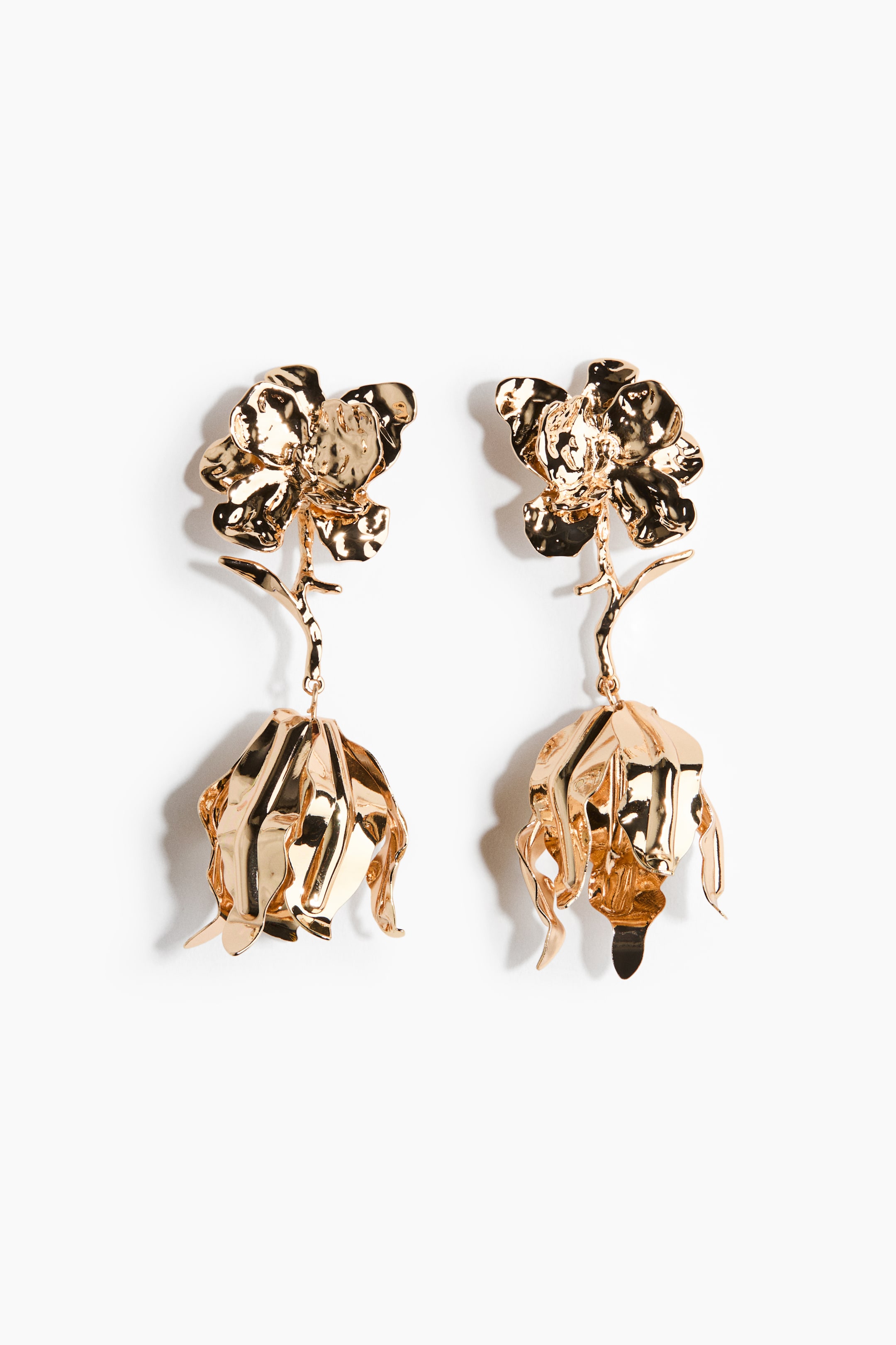 Visa större bild: Ett par guldfärgade hängande örhängen med en hamrad blomdesign. Varje örhänge visar en texturerad öppen blomma upptill, som hänger en klockformad, hängande blomknoppselement nedan med en böjd stjälk.