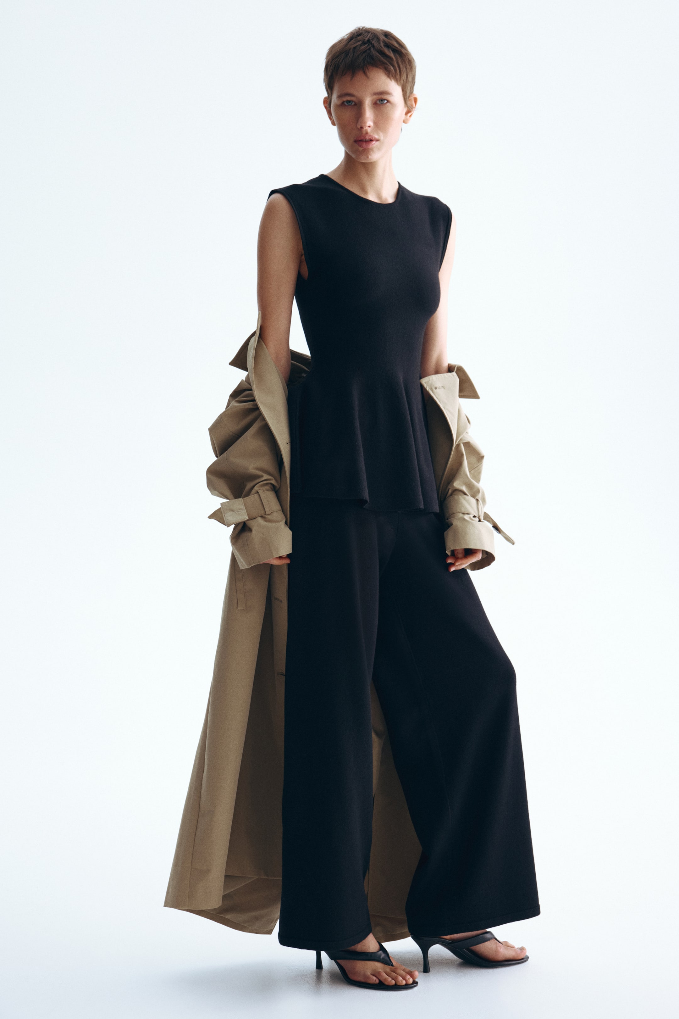 View larger image: A black sleeveless top featuring a round neck and a peplum hem is paired with black wide-leg trousers. A beige trench coat is draped over the shoulders, complemented by black thong-style high-heeled sandals.
