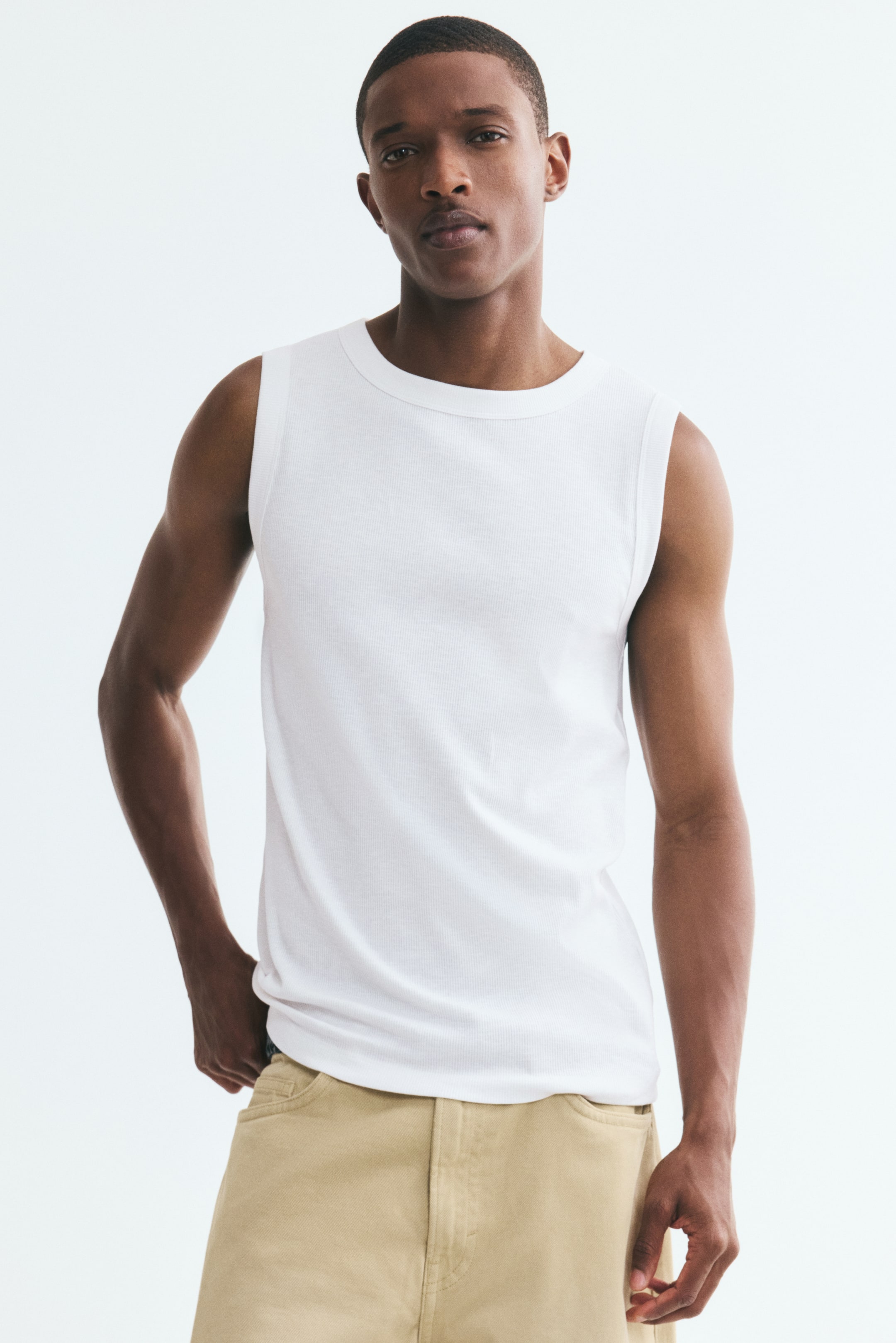 View larger image: The model is wearing a white vest top in a ribbed cotton-blend jersey, featuring a round neckline, sleeveless design, and slim, form-fitting silhouette. The top is styled with beige trousers.
