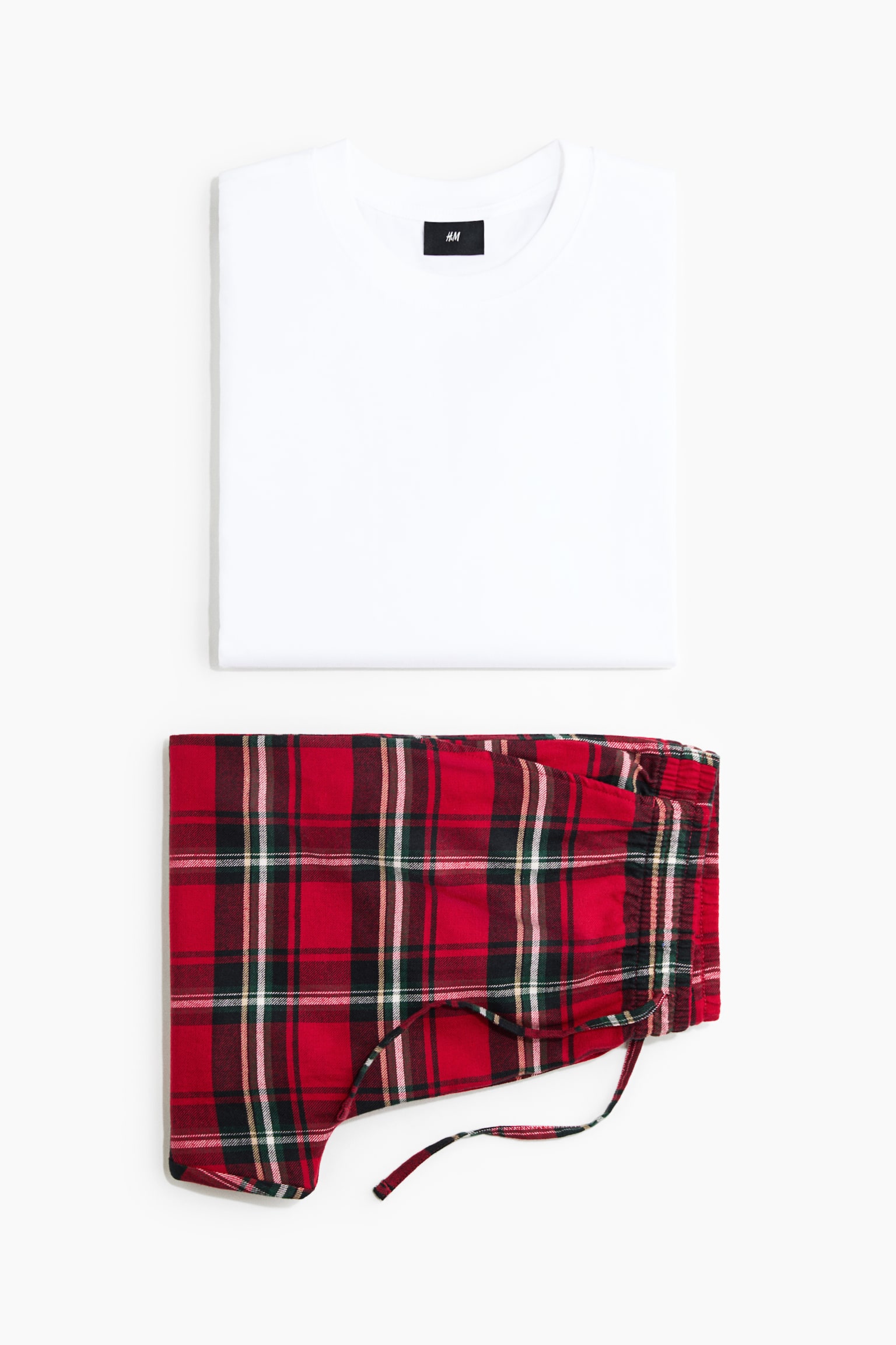 Ein Pyjama-Set ist in einer frontalen Flatlay-Ansicht zu sehen und zeigt ein zusammengelegtes weiÃes Jersey-Oberteil aus Baumwolle mit Rundhalsausschnitt, das Ã¼ber zusammengelegten roten Flanellhosen mit Karomuster platziert ist. Die Hose ist mit einem elastischen Bund und einem passenden Kordelzug im Karomuster dargestellt.