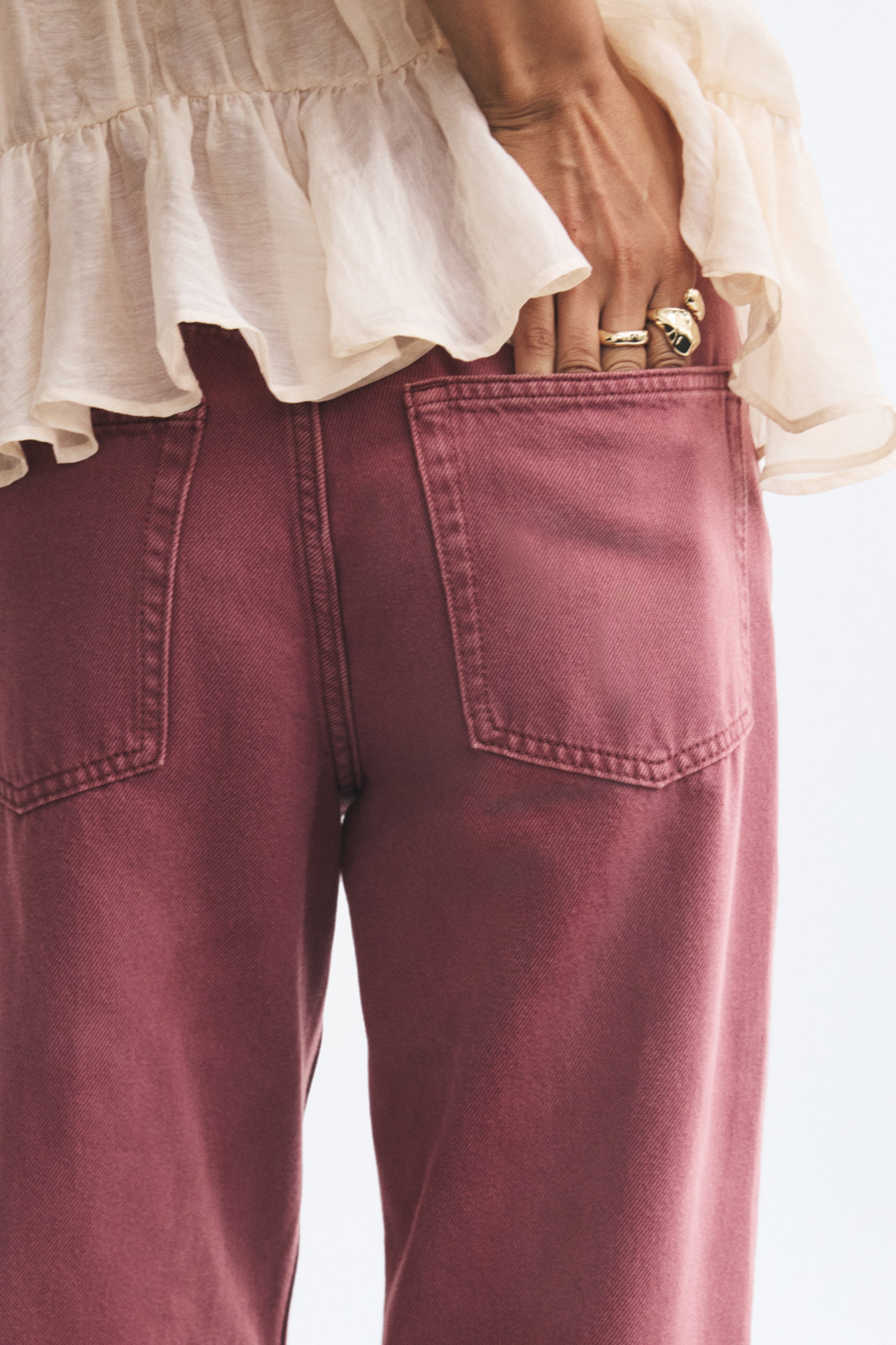 View larger image: The model is wearing muted burgundy denim jeans, sliding a hand into the right back pocket. The jeans feature visible stitching on the rear patch pockets and are paired with a ruffled, semi-sheer cream blouse.