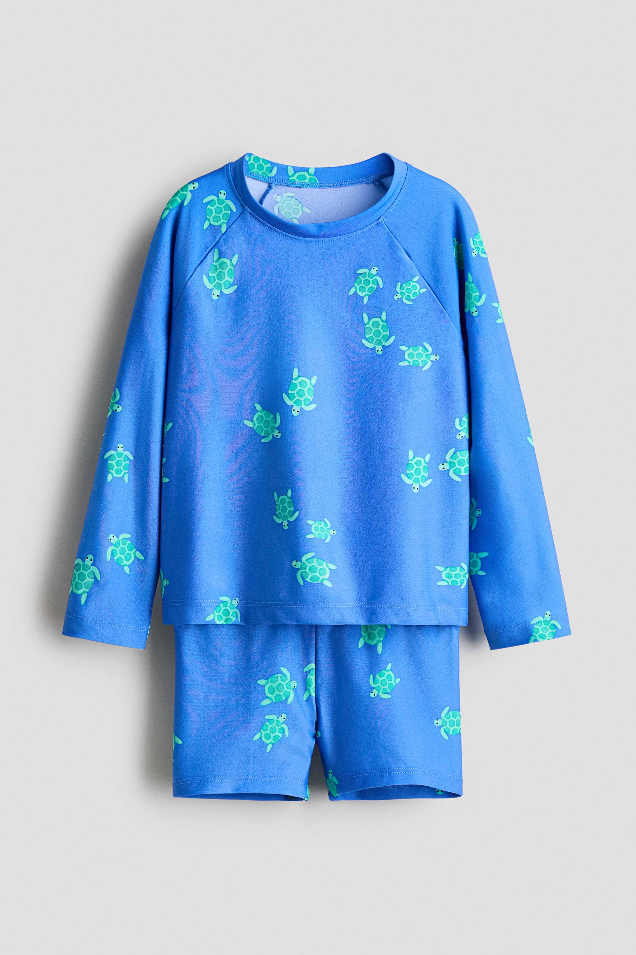 Ver imagem maior: Um conjunto infantil azul de rash guard apresenta uma peça superior de mangas compridas com crew neck e mangas raglan, e calções combinantes, ambos cobertos por um padrão repetido de tartarugas verdes, voltadas para a frente.