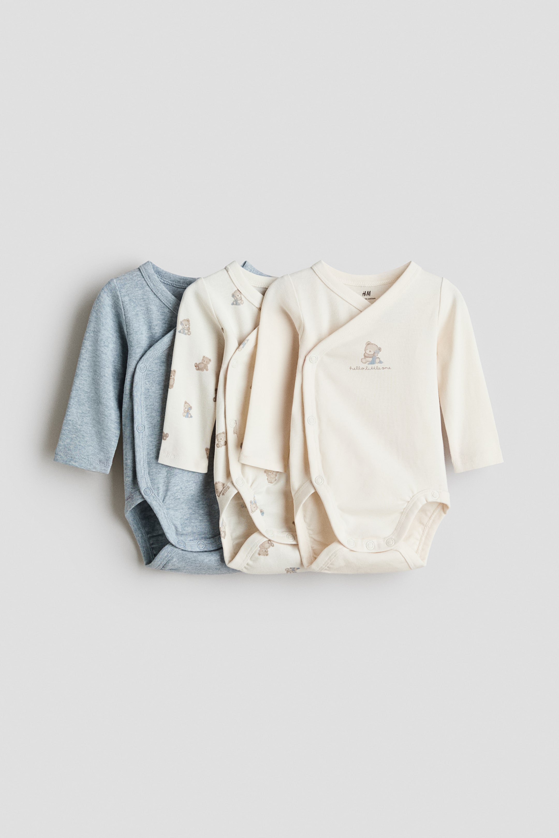 Visa större bild: Tre långärmade babybodys är lagda i lager, vända framåt och något åt höger. Den främre krämfärgade bodyn har en omlottstil med sid- och grenknappar, ett broderat teddybjörnsmotiv och texten "hello little one". Den mellersta krämfärgade bodyn visar ett heltäckande teddybjörnsmönster, och den bakre blå bodyn är designad med rund hals och grenknappar.