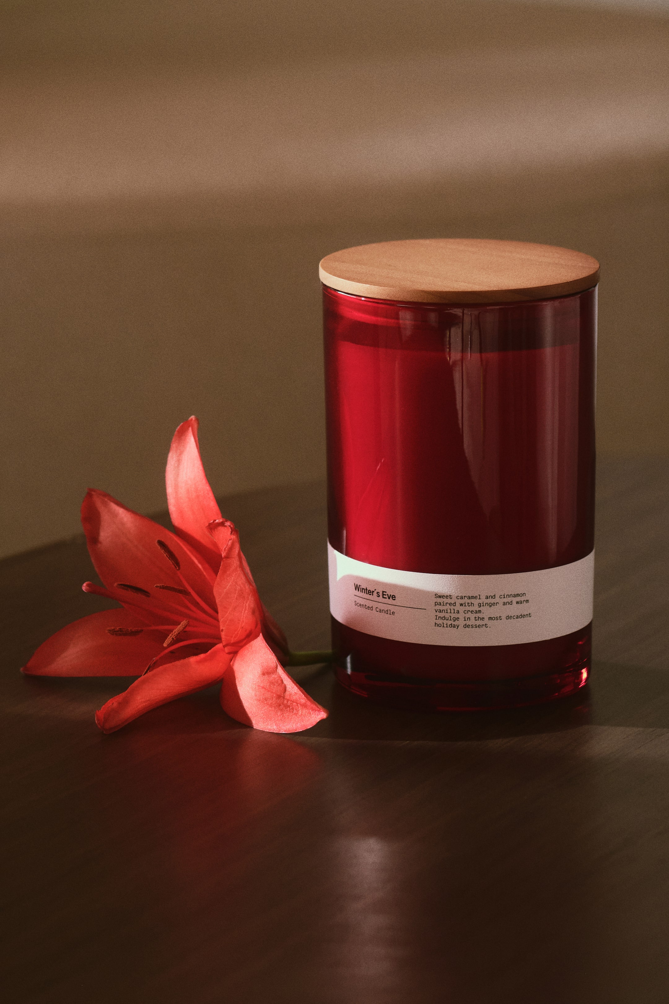 Ver imagen más grande: Una vela cilíndrica perfumada en un portavelas de vidrio rojo intenso se mantiene en pie con una tapa plana de madera apoyada encima. Una etiqueta de papel blanca rodea la parte inferior del vidrio, y un único lirio rojo yace sobre la superficie junto a la vela.