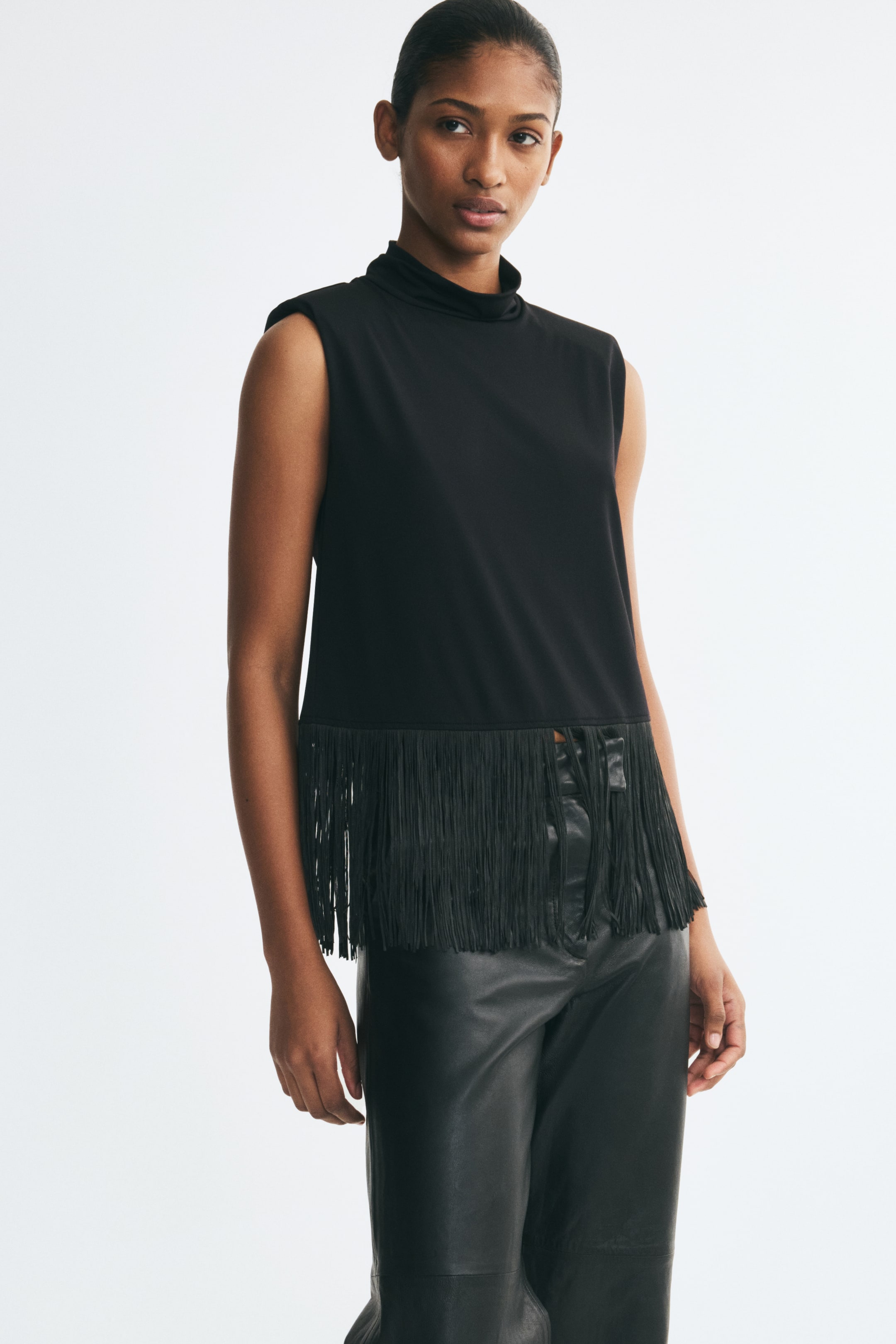 View larger image: The model is wearing a sleeveless black top with a high neck, structured shoulder pads, and long fringe at the hem, paired with black leather trousers.