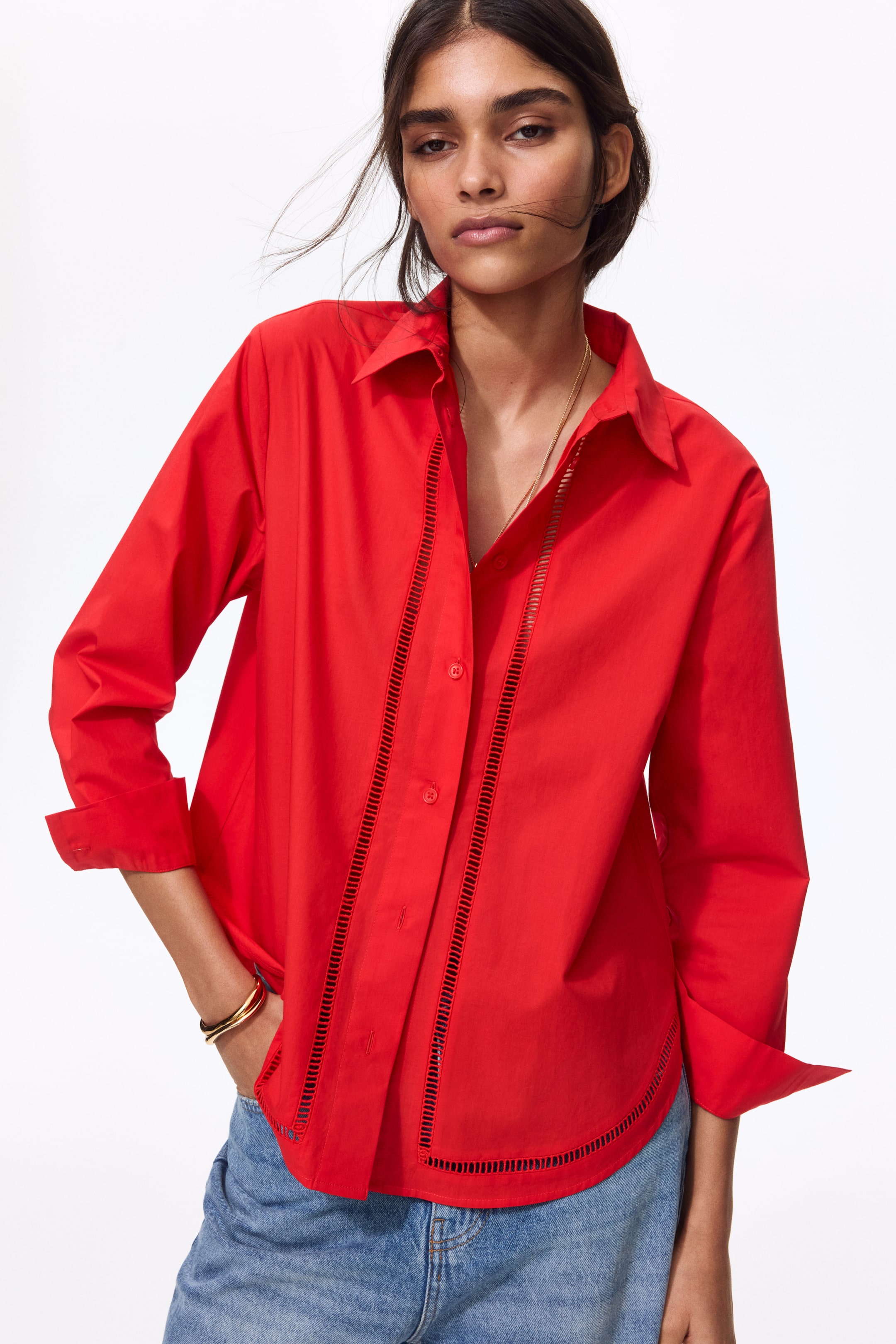 View larger image: A crisp red cotton poplin shirt, showcasing ladder lace insets down the front and along its rounded hem, is being worn by the model with its sleeves rolled up. The shirt is also featuring a collar and buttons down the front, and the model is styling it with blue jeans.