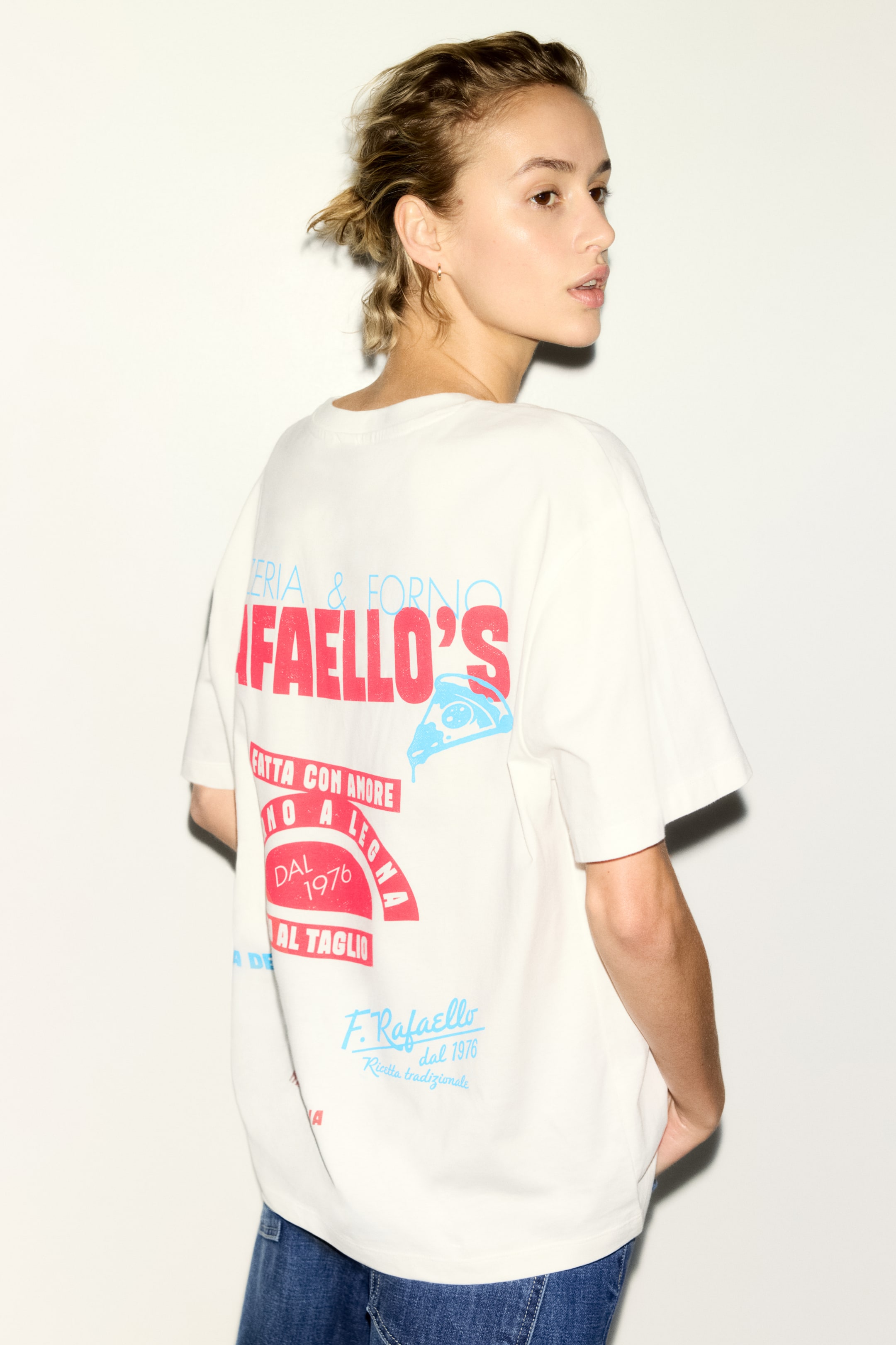 View larger image: An off-white, loose-fitting t-shirt is being viewed from the back, showcasing a multi-coloured graphic print. The print is featuring "Rafaello's" in prominent red, along with other text in red and light blue, and a light blue stylized pizza slice graphic with a bite taken out and dripping, while its short sleeves are falling loosely.