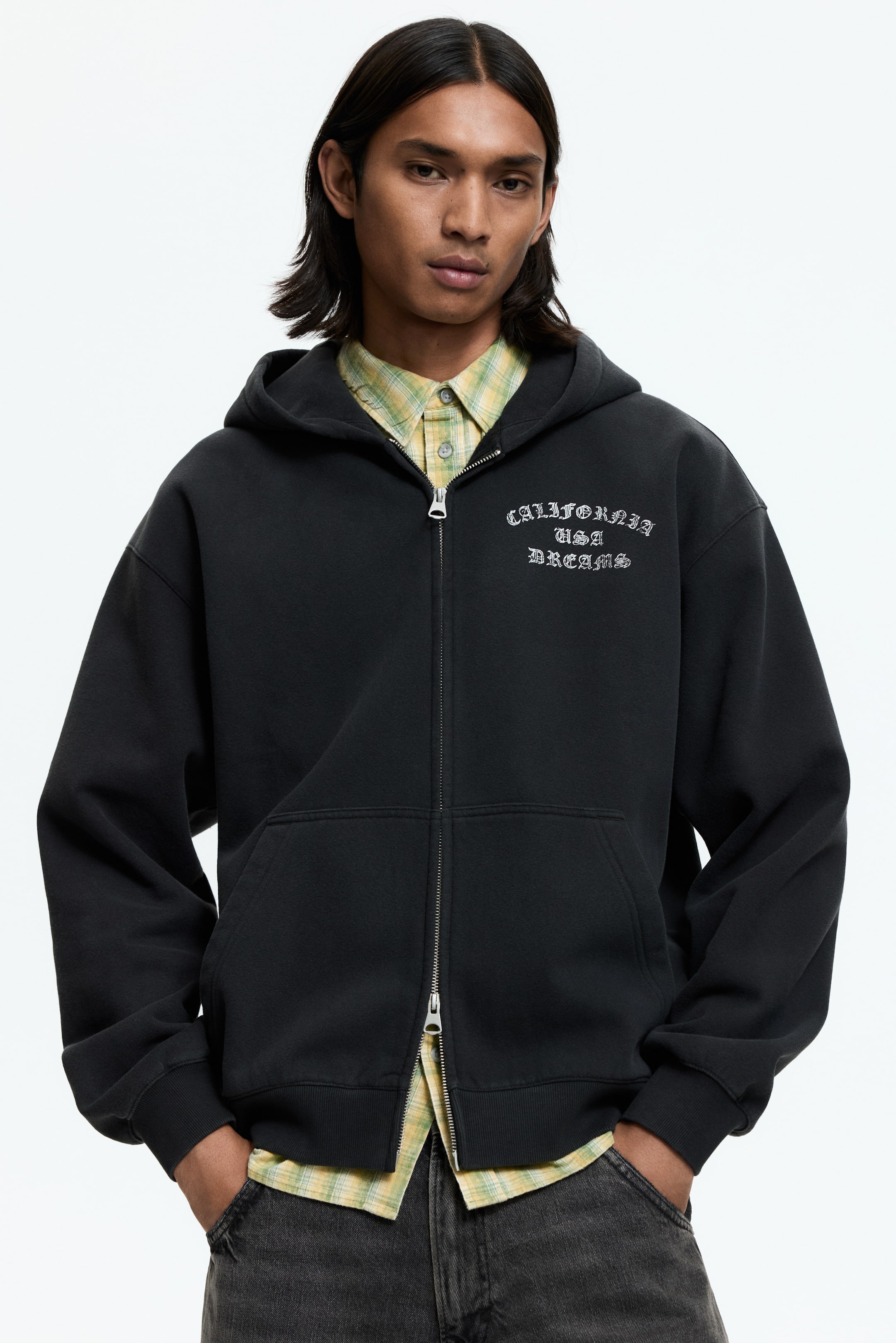 Größeres Bild ansehen: Ein schwarzer Kapuzenpullover mit Reißverschluss zeigt sich von vorne, ausgestattet mit einem durchgehenden metallischen Reißverschluss, Kängurutaschen, gerippten Bündchen und einem gerippten Saum. Auf der Brust ist in Weiß der gestickte Text "CALIFORNIA USA DREAMS" zu sehen, über einem hellgelben karierten Hemd.