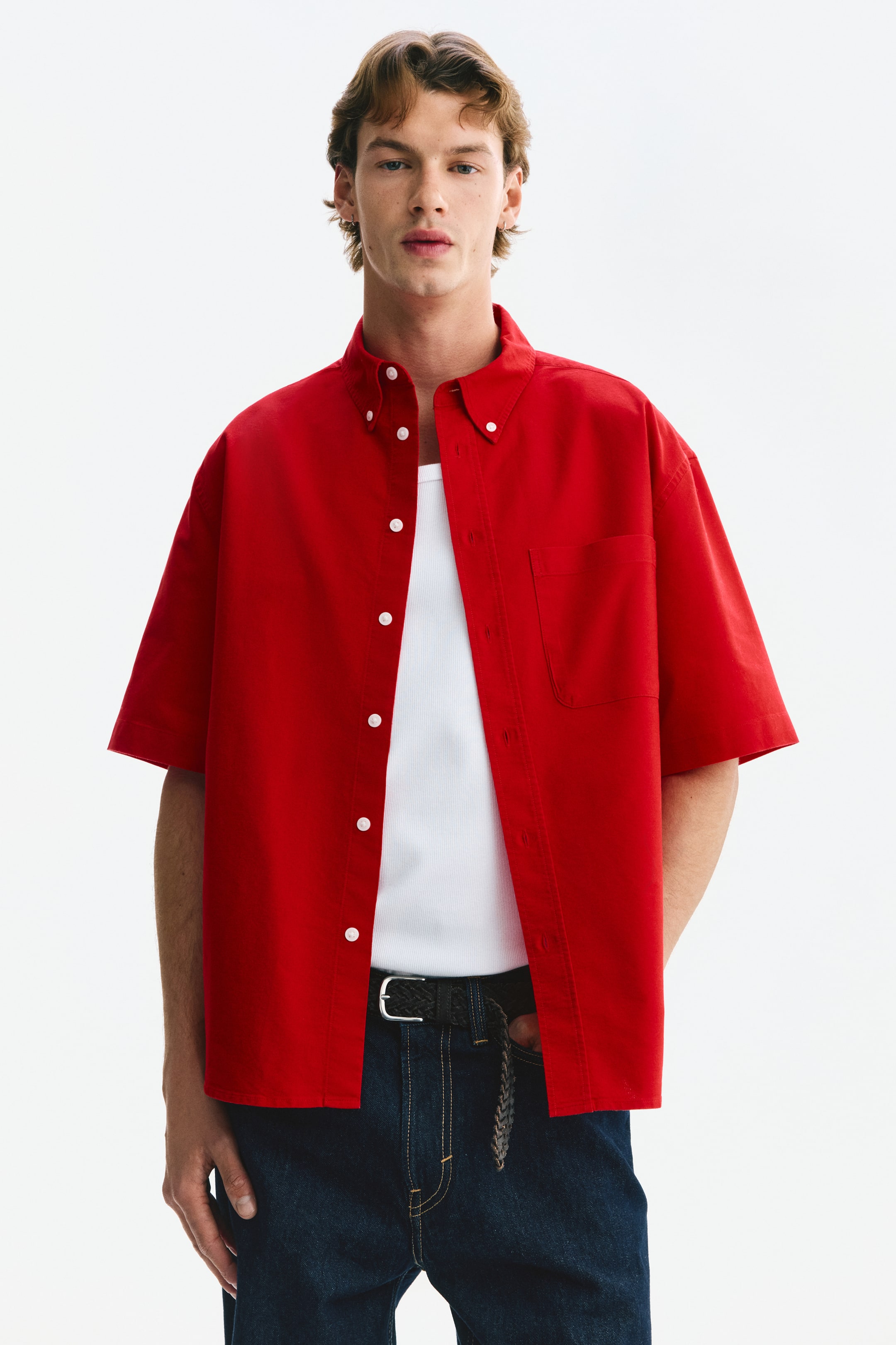 View larger image: Presenting a red short-sleeved button-front shirt, designed with a button-down collar, showcasing white buttons along the front and a patch pocket on the left chest. The shirt is styled open over a white undershirt.