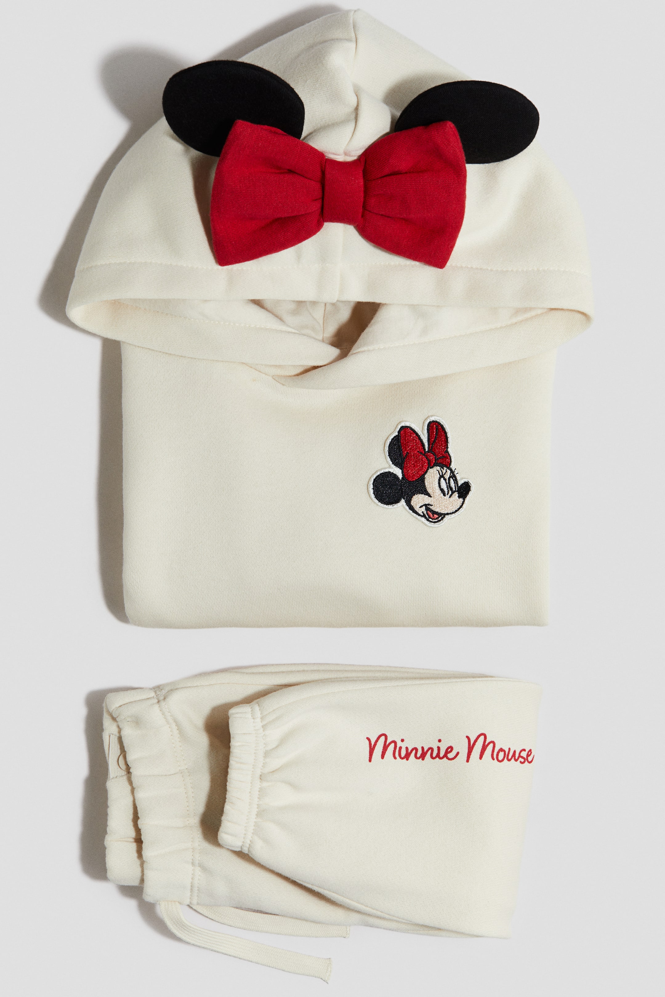 Zobraziť väčší obrázok: Krémovo sfarbená dvojdielna súprava na voľný čas je vystavená, obsahuje mikinu s kapucňou smerujúcou dopredu s čiernymi kruhovými ušami a červenou mašľou na kapucni a vyšívanou hlavou Minnie Mouse na ľavej strane hrudníka. Zodpovedajúce teplákové nohavice majú elastické členky a červený vyšívaný text "Minnie Mouse" na jednej nohe.