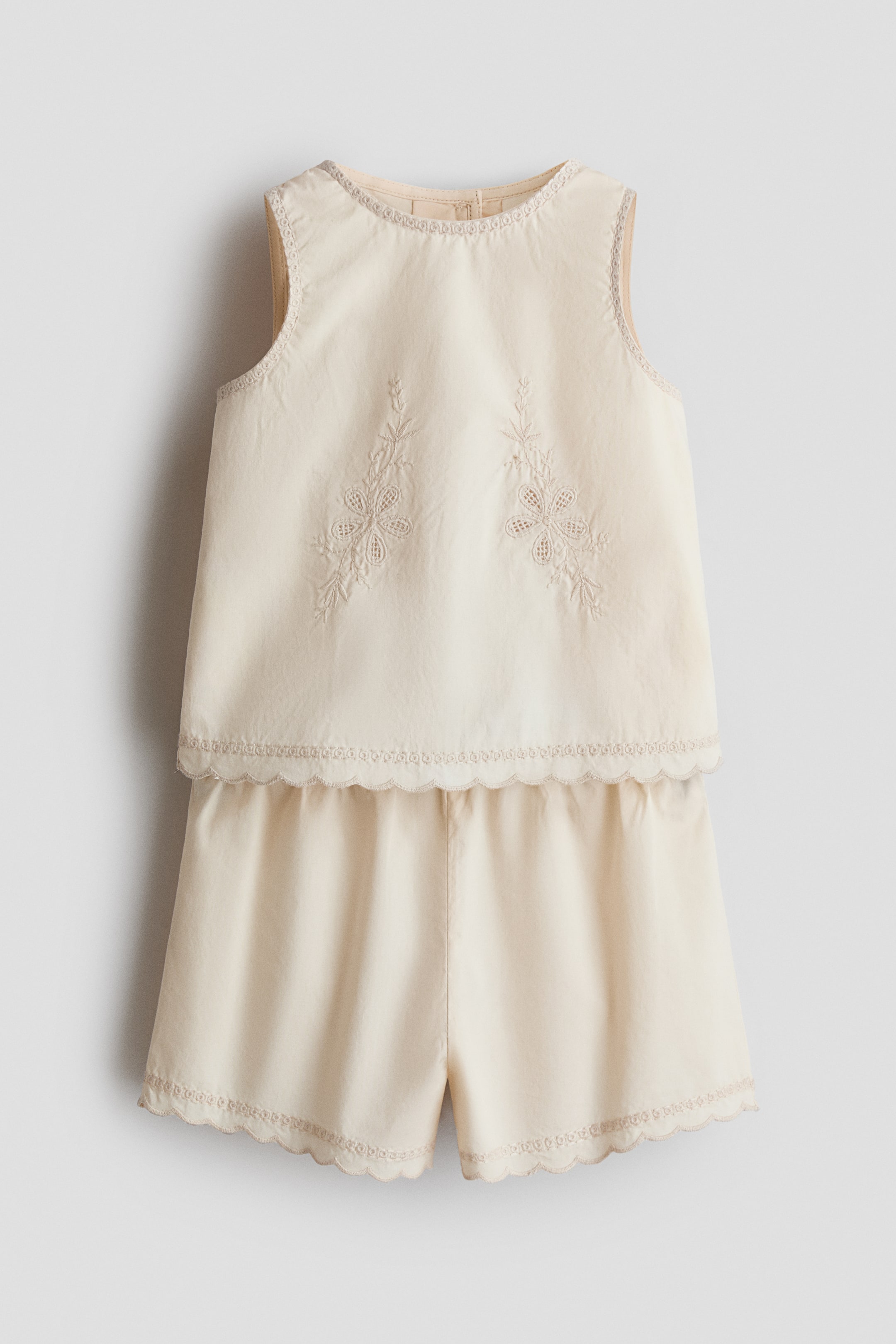 View larger image: Presenting a cream two-piece set from the front, this ensemble features a sleeveless top with a round neckline, adorned with two embroidered floral patterns on the front. The top is detailed with delicate scalloped trim along the neckline, armholes, and bottom hem. This same elegant trim also borders the leg openings of the matching shorts.