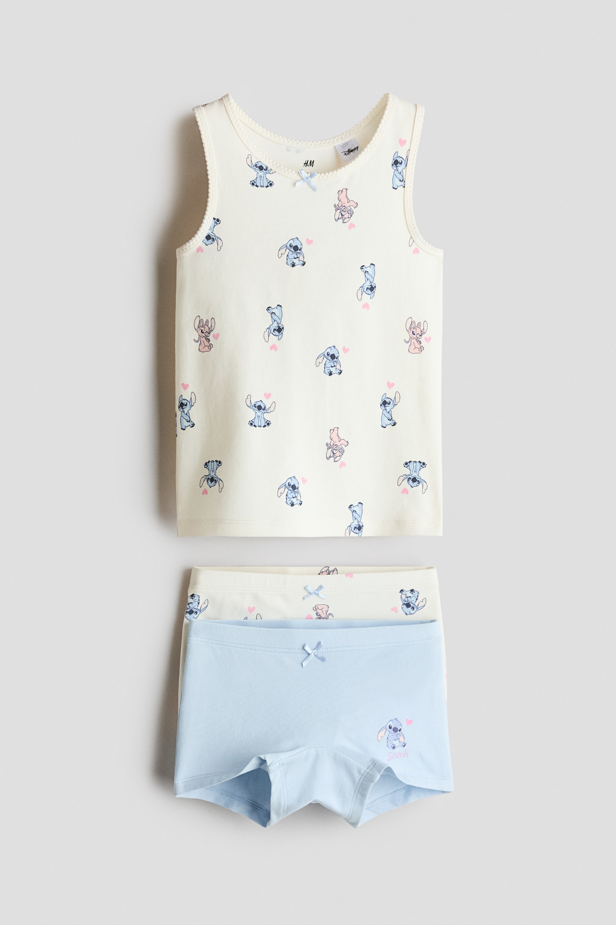 Grösseres Bild ansehen: Ein Set präsentiert ein cremefarbenes Unterhemd, das mit einem Allover-Print der Figuren Stitch und Angel, einer Picot-Borte entlang des Ausschnitts und der Armlöcher sowie einer kleinen Schleife vorne in der Mitte versehen ist. Alles wird von vorne präsentiert und flach hingelegt. Darunter sind zwei Slips geschichtet, wobei der obere hellblaue Slip einen einzelnen Stitch-Print und eine kleine Schleife zeigt, und der untere cremefarbene Slip einen Allover-Print der Figuren Stitch und Angel sowie eine kleine Schleife aufweist.