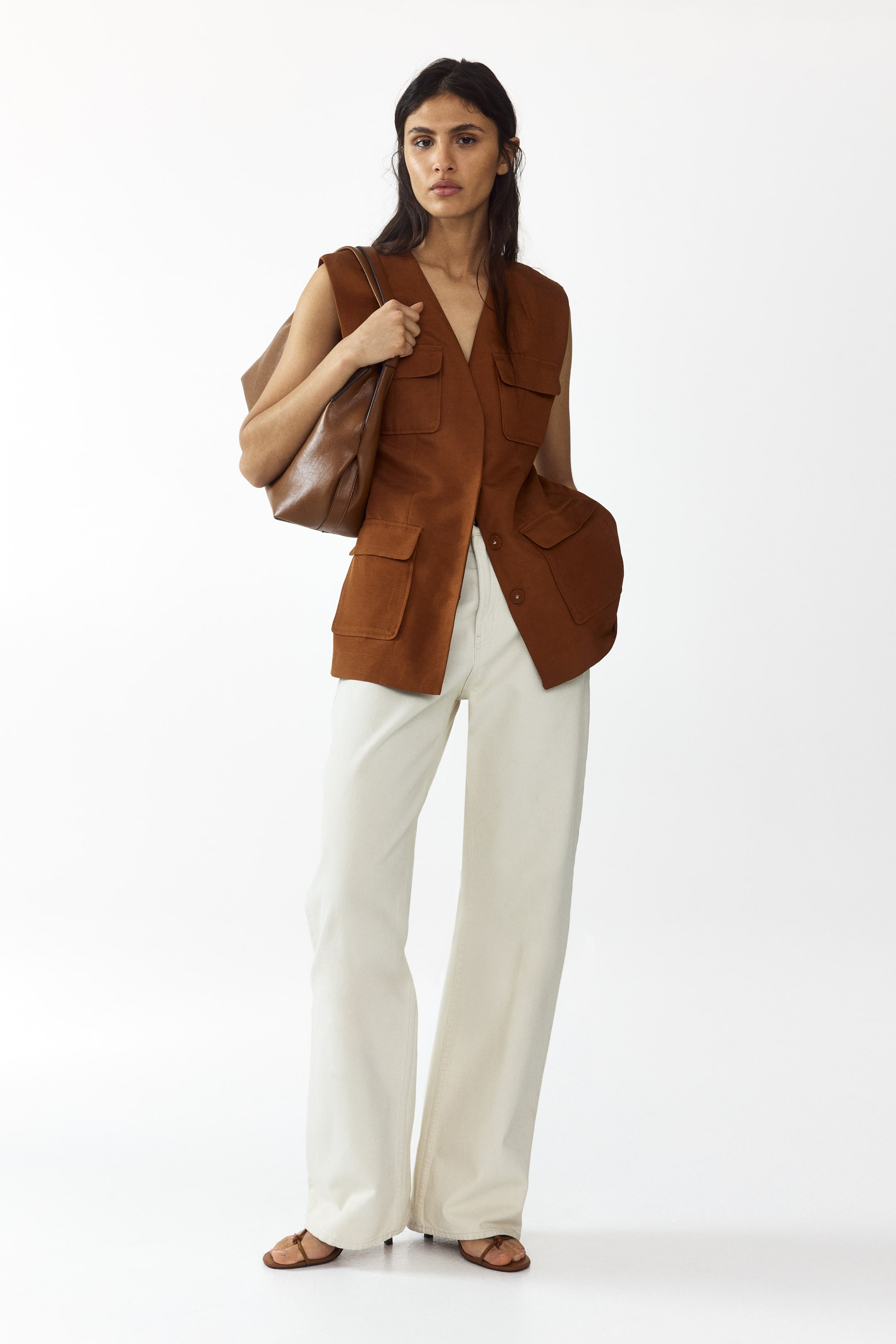 Prikaži veću sliku: Model nosi pripijeni prsluk bez rukava u boji rđe, koji ima V-izrez, zatvoreni prednji deo i četiri džepa s preklopom. Model stavlja jednu ruku u donji prednji džep i nosi smeđu torbu preko ramena, dok prsluk kombinuje sa svetlim pantalonama širokih nogavica.