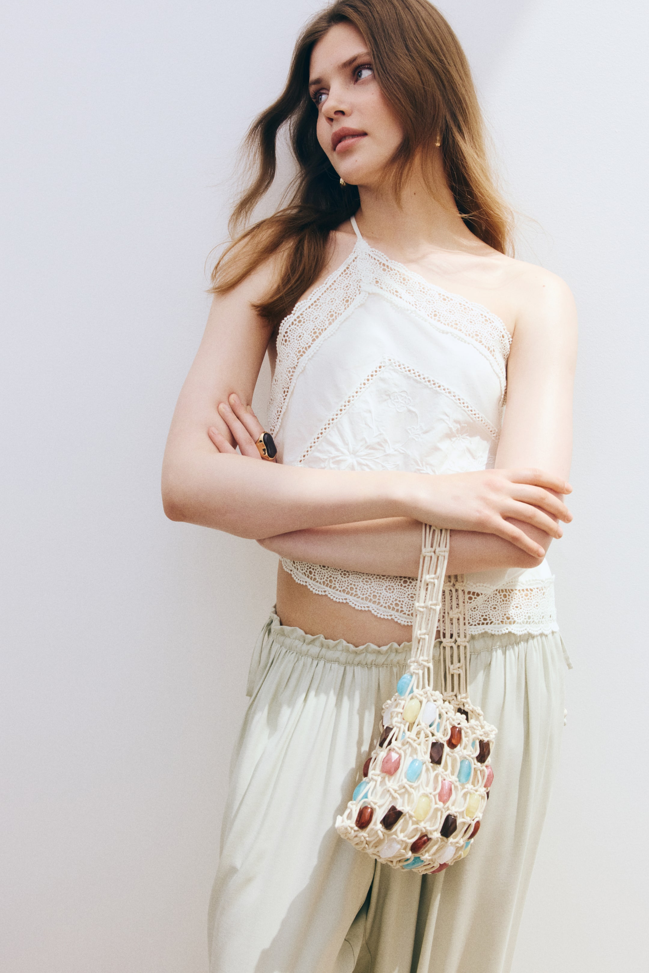 Grotere afbeelding bekijken: Een witte halter crop top met verfijnde kanten afwerking langs de halslijn en zoom, met delicate borduursels zichtbaar op het hoofdgedeelte. Het wordt gecombineerd met lichtgroene wijde broekspijpen, die een geplooide taille en een losse val vertonen.