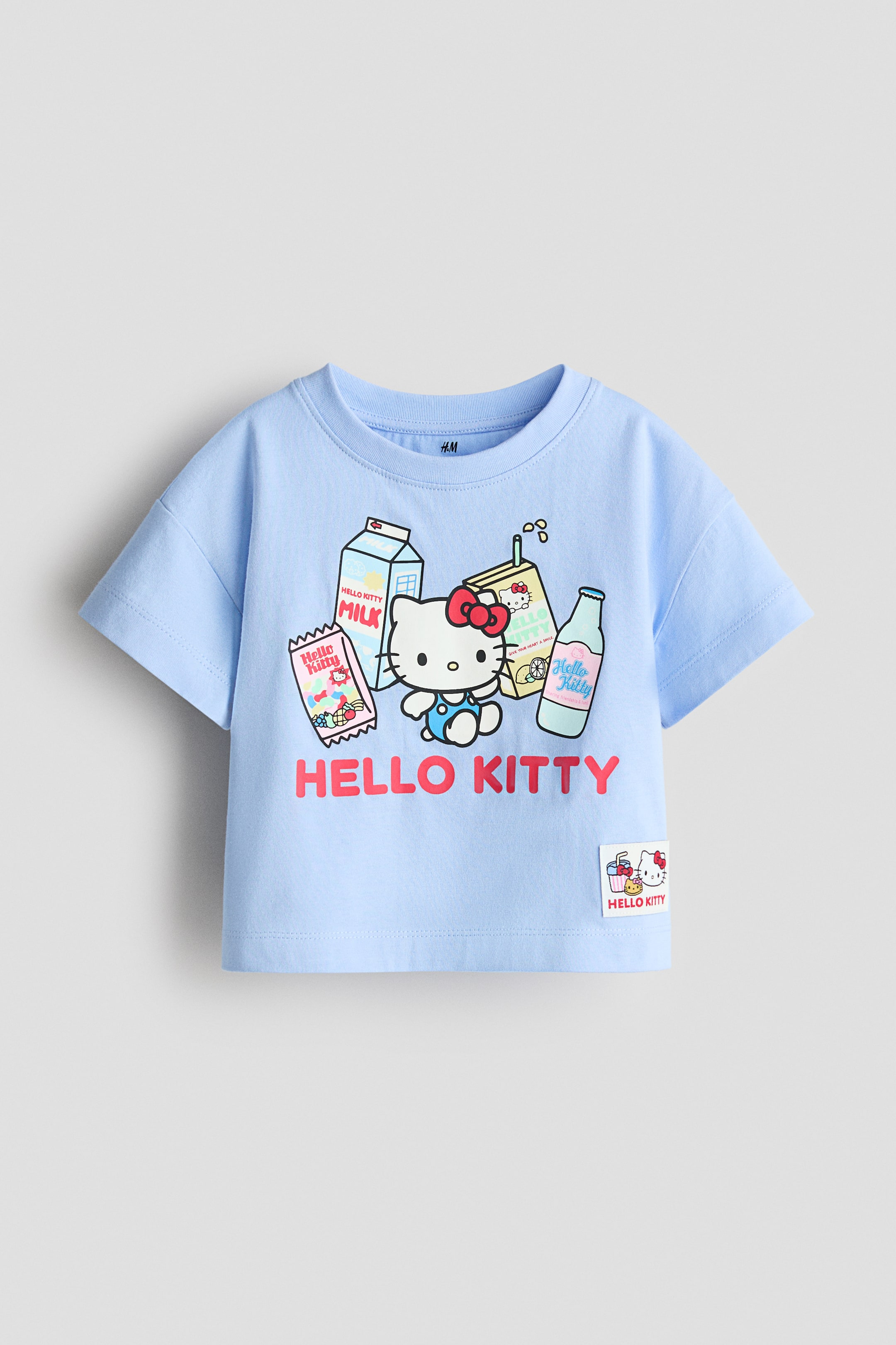 View larger image: A light blue short-sleeved t-shirt is presenting a crew neck and a colorful graphic on its front. The graphic features Hello Kitty wearing blue overalls, surrounded by branded milk cartons, a juice box, a candy wrapper, and a bottle, with "HELLO KITTY" text in red below. A small woven label showing a Hello Kitty graphic is also visible on the bottom right hem.