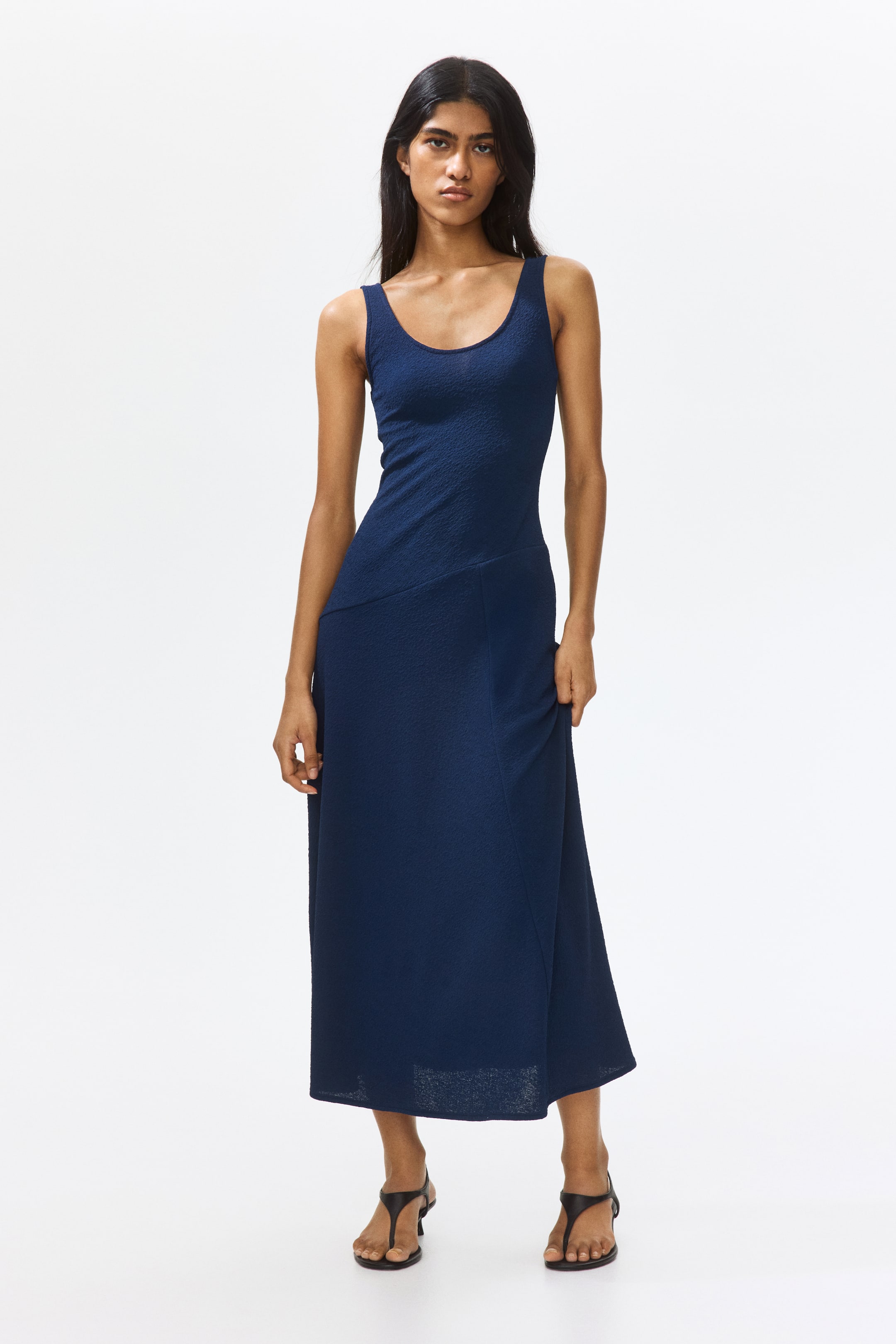 View larger image: The model is wearing an ankle-length, sleeveless dress crafted from dark blue jersey that has a subtly felted appearance. The dress features a scoop neckline, a diagonal seam at the hips, and a flared skirt that reveals a high slit, with the model gently holding the fabric of the skirt.