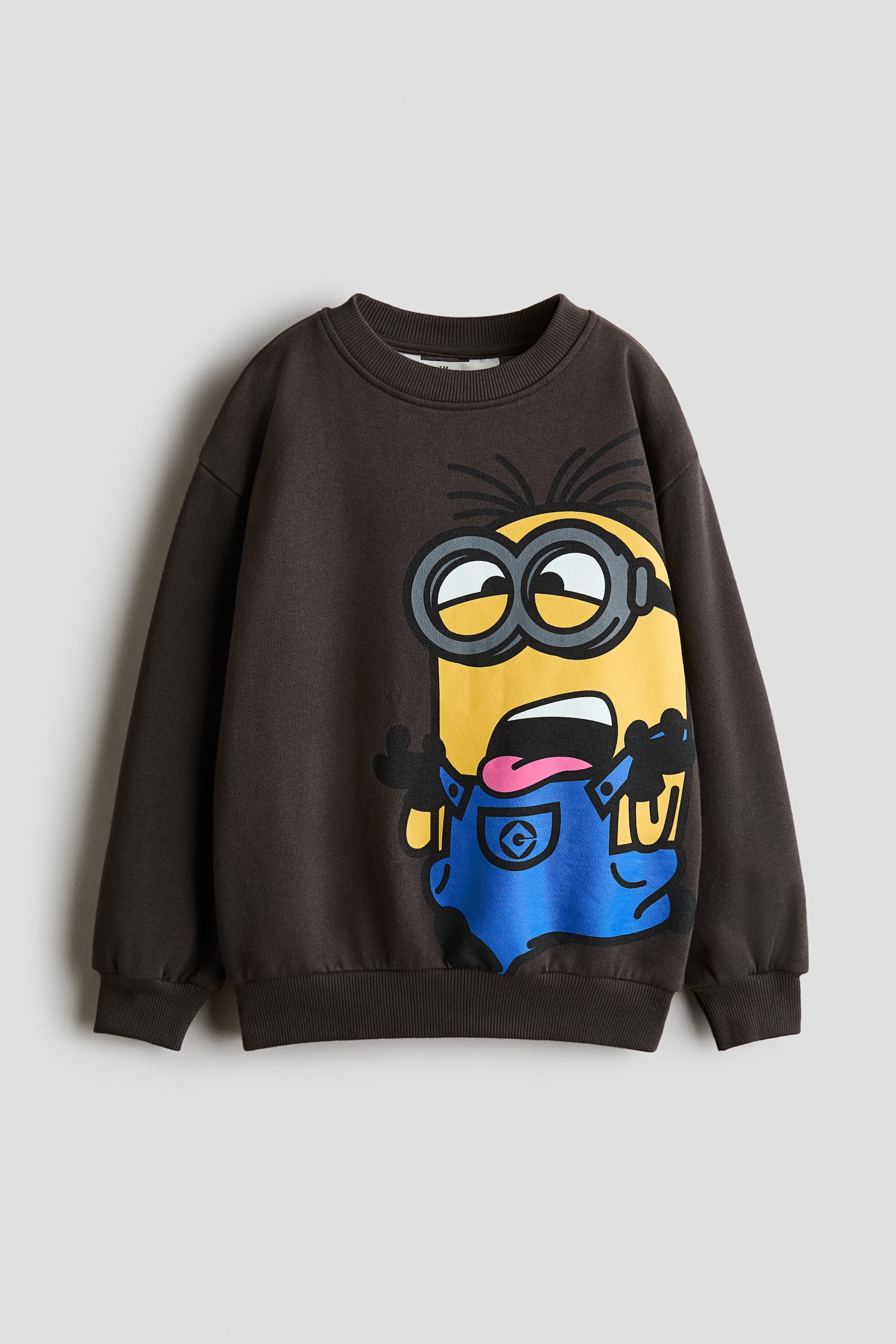 En mÃ¸rkebrun sweatshirt vises forfra og fremhÃ¦ver et stort grafisk print af en Minion-figur i blÃ¥ overalls pÃ¥ hÃ¸jre side. Plagget har en ribstrikket rund halsudskÃ¦ring, sÃ¦nkede skuldre samt ribstrikkede kanter ved manchetterne og nederkanten.