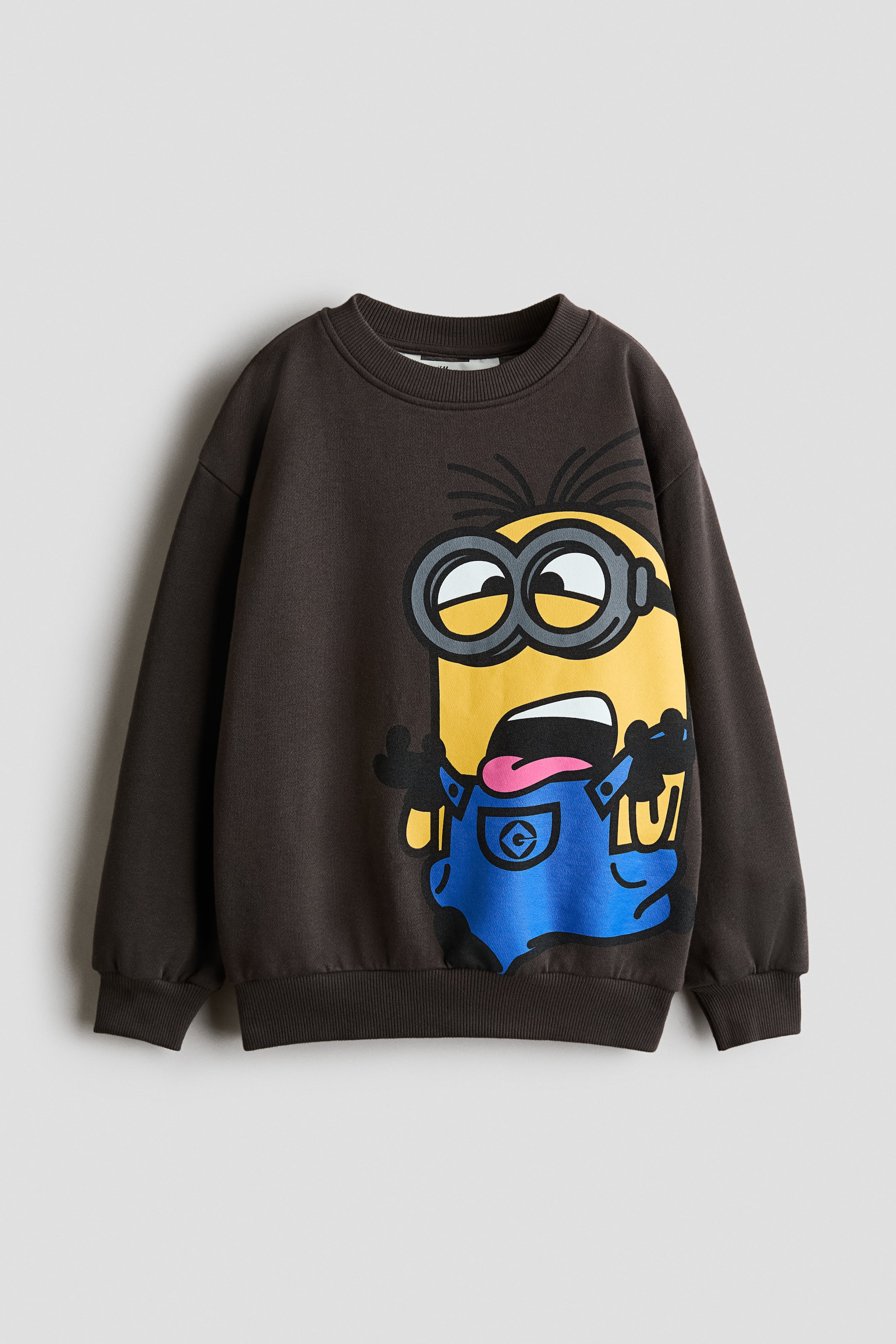 Agrandir l'image: Un sweat-shirt marron foncé est présenté de face, mettant en valeur un grand motif imprimé représentant un personnage Minion en salopette bleue sur le côté droit. Le vêtement présente un col rond côtelé, des épaules tombantes, ainsi que des finitions côtelées aux poignets et à l’ourlet.