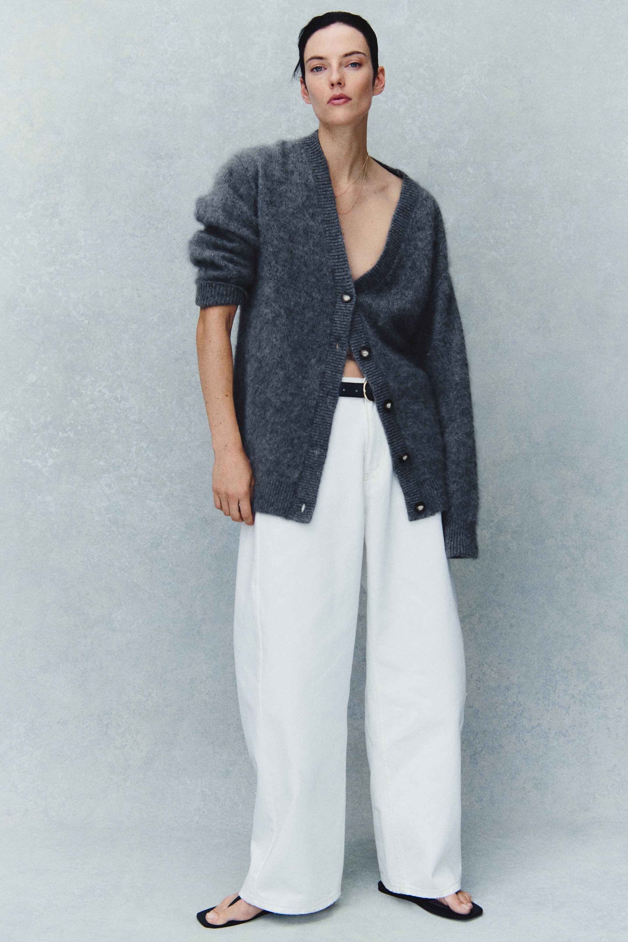 View larger image: An oversized gray cardigan in a brushed wool knit is worn by the model, styled partially buttoned over white wide-leg trousers. The cardigan features a deep V-neckline, dropped shoulders with one sleeve pushed up, and ribbed detailing along the neckline, button band, cuffs, and hem.