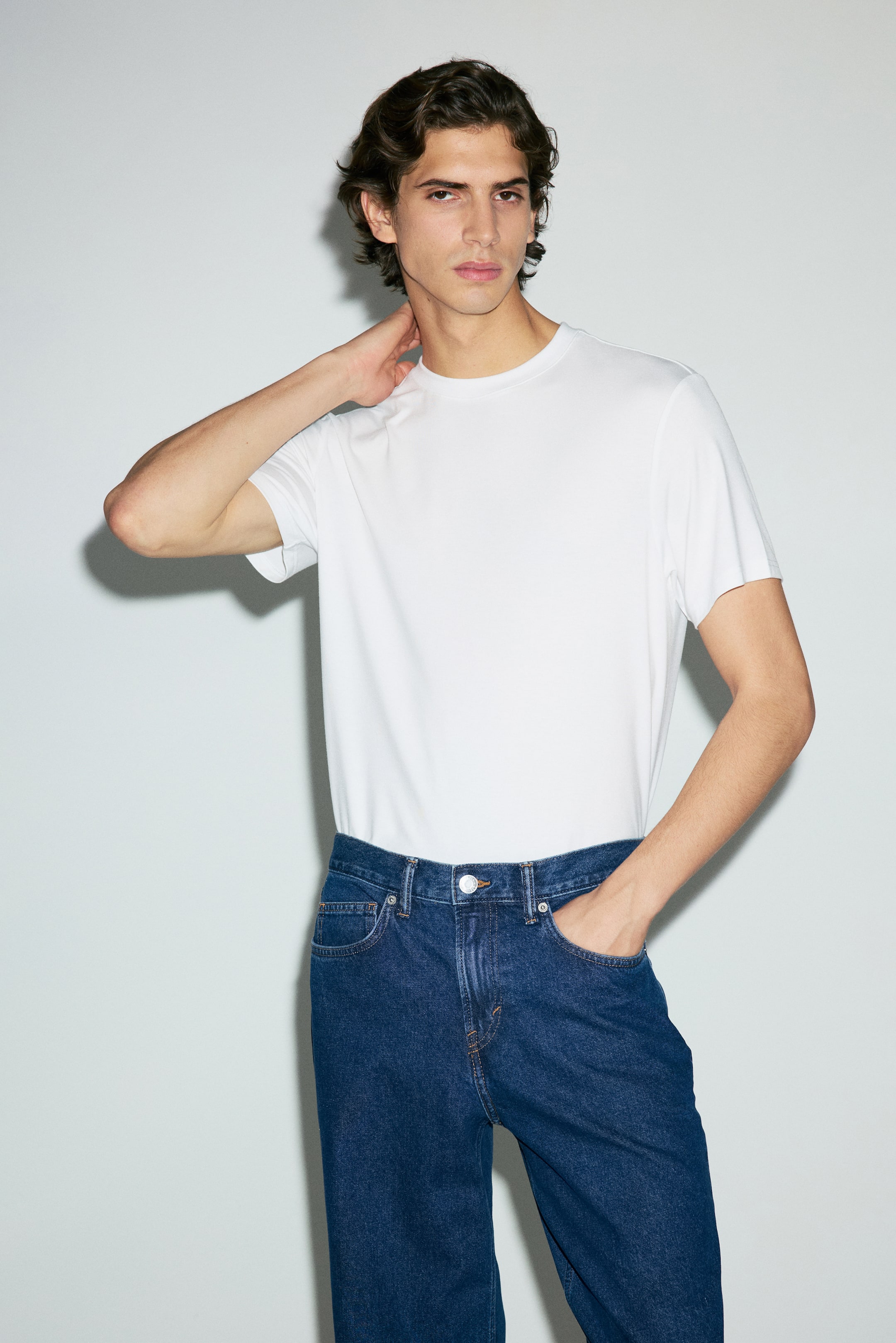 Se større billede: En hvid t-shirt med korte ærmer og rund hals bæres, stukket ned i mørkvaskede denimjeans. Jeansene har en sølvfarvet knaplukning, synlige forlommer og let kontrastsyning, alt præsenteret fra en let vinklet frontvisning.