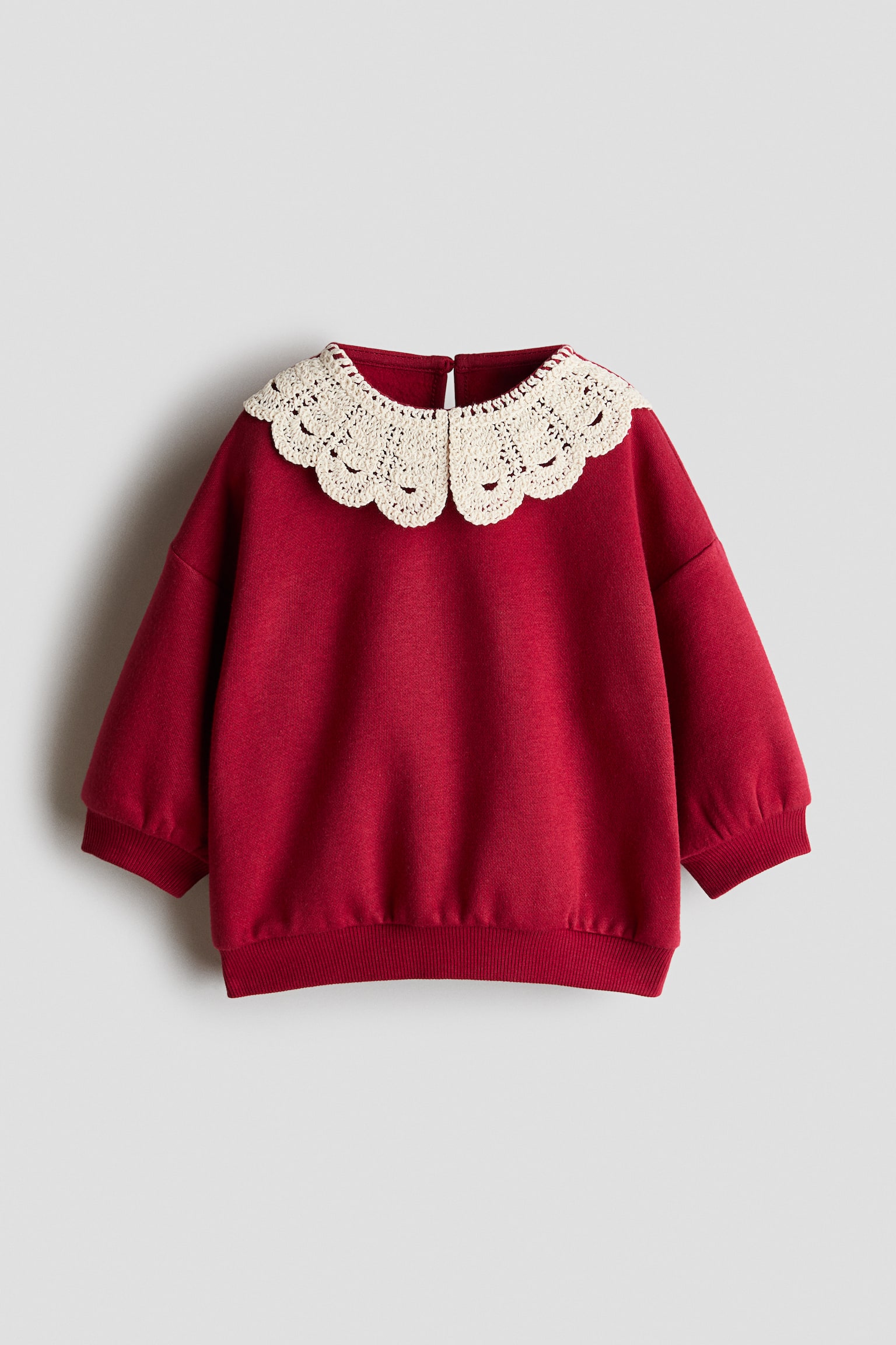 Una sudadera roja vista de frente muestra un gran cuello de crochet color crema con borde ondulado. El suÃ©ter tipo pulÃ³ver presenta hombros caÃ­dos, puÃ±os acanalados y dobladillo acanalado.