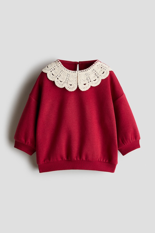 A red sweatshirt shown from the front features a large, cream-coloured scalloped crochet collar. The pullover has drop shoulders, ribbed cuffs, and a ribbed hem. - 1