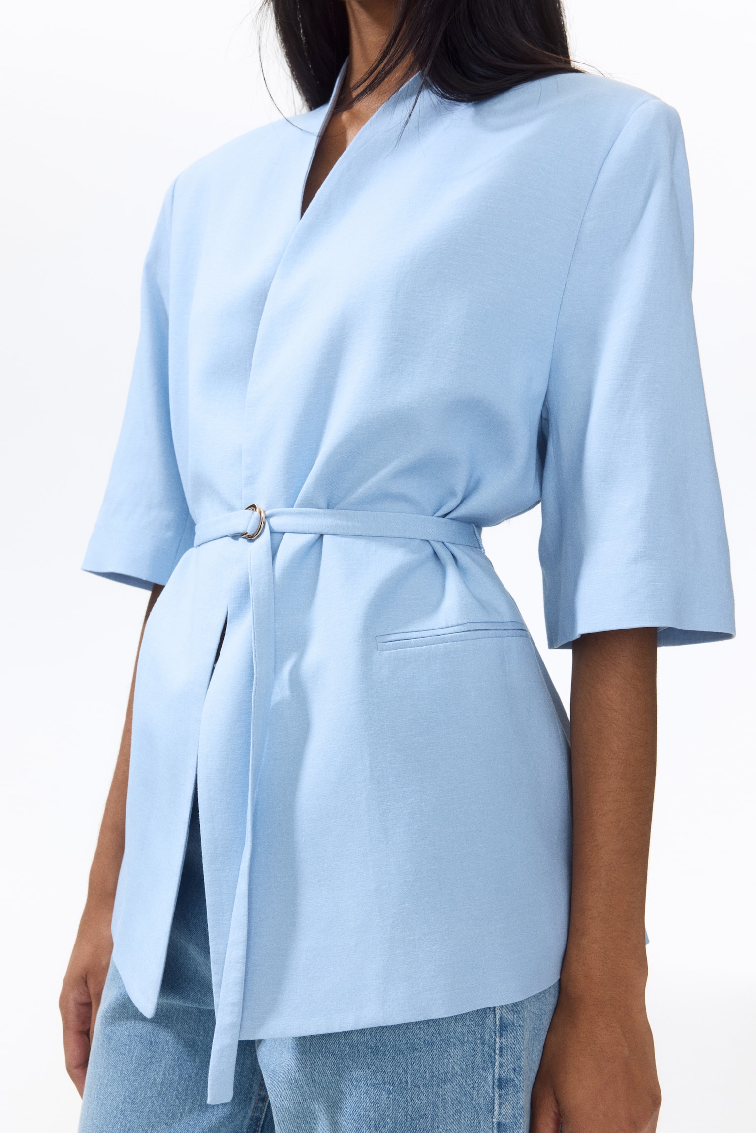A light blue wrap jacket is showcased from a front-side angle, featuring a collarless V-neck and wide elbow-length sleeves. A self-fabric belt cinches the waist with a metallic D-ring buckle, and a welt pocket is visible on the right side. - 1