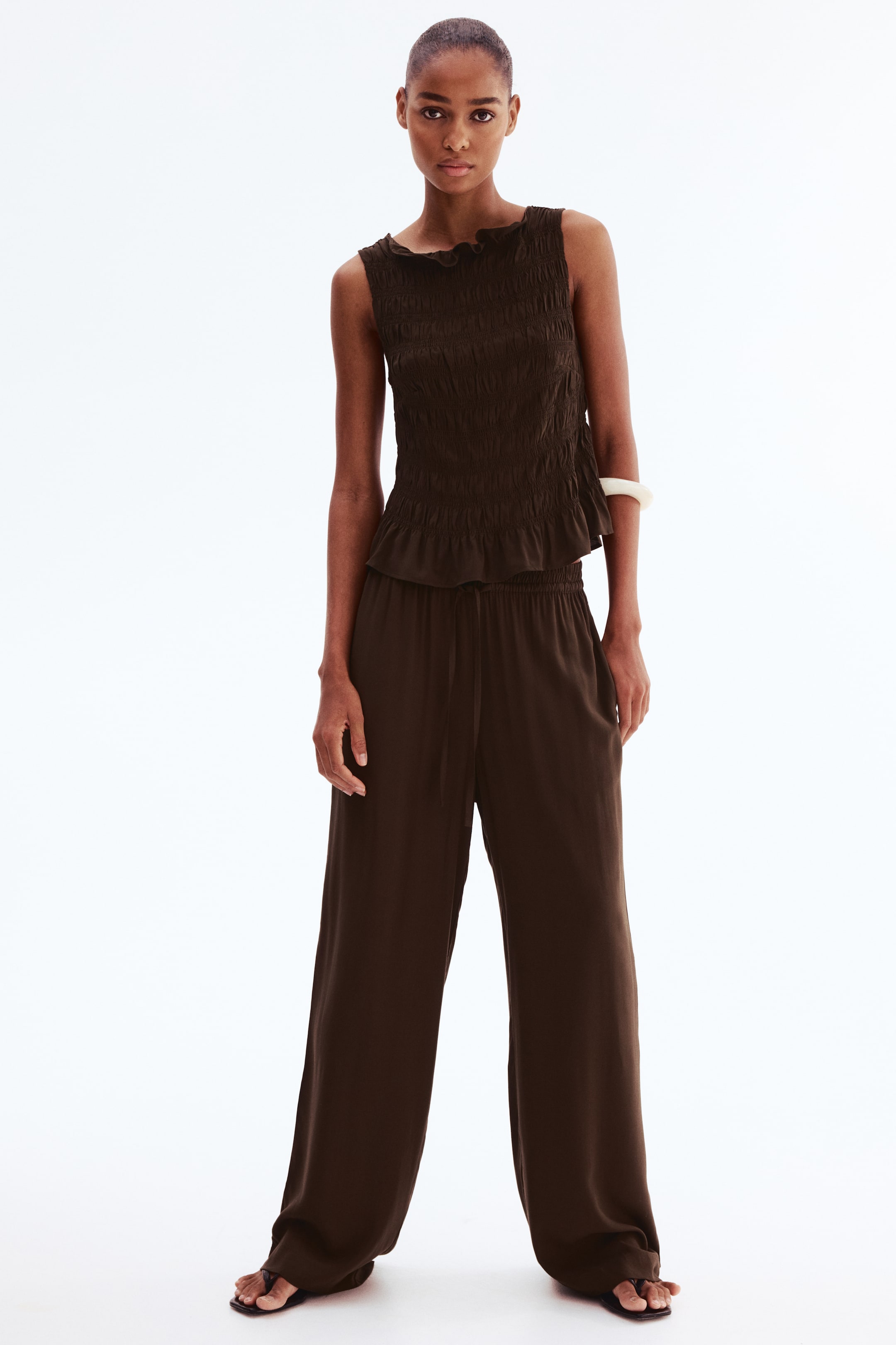 View larger image: Loose-fitting dark brown pants in a viscose weave are worn by the model, featuring a high waist with a drawstring and wide legs. The model’s hands rest near the discreet side pockets, and the pants are styled with a matching sleeveless top.