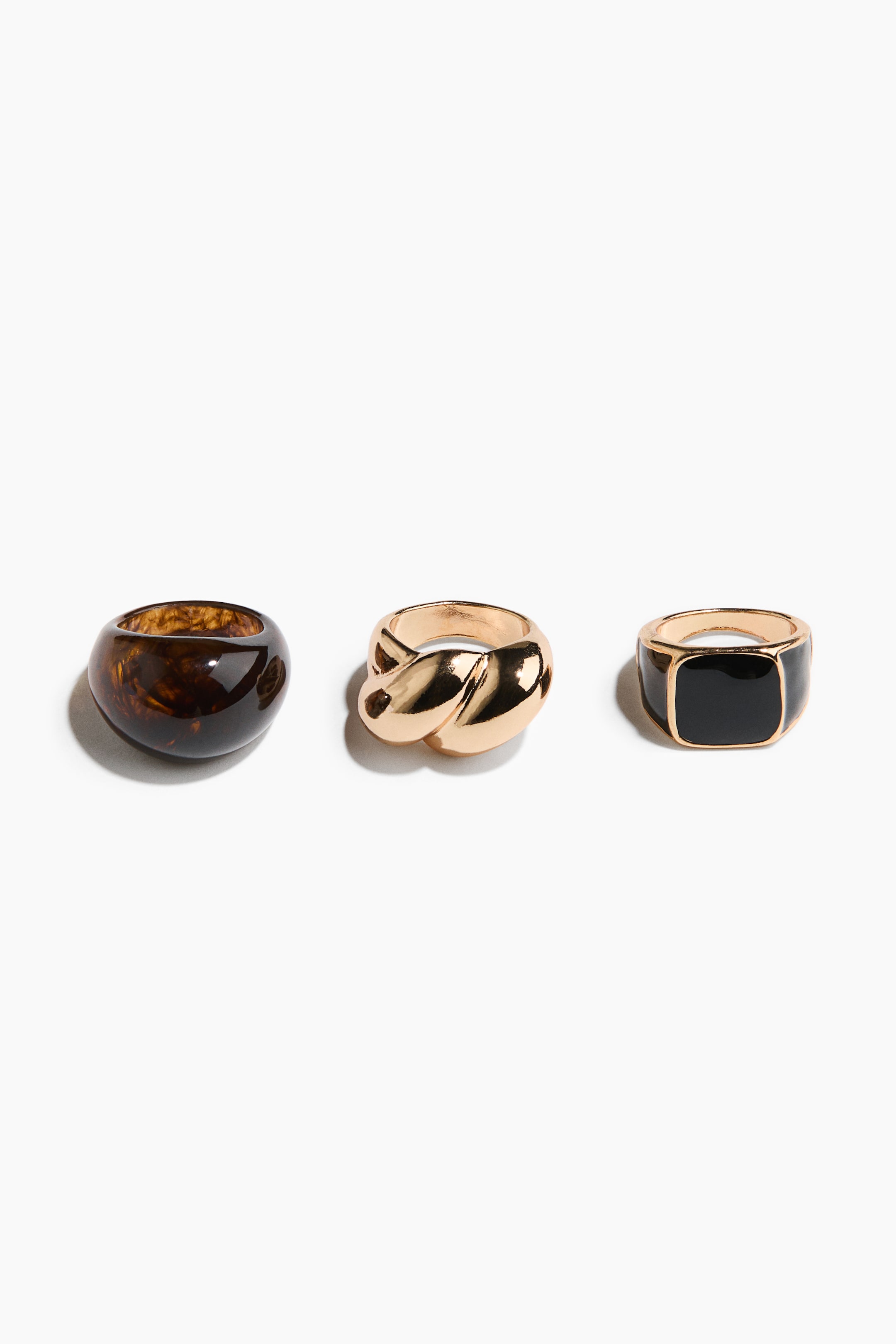 View larger image: Three chunky rings showcase diverse designs, each angled slightly to the right. On the left sits a rounded tortoiseshell resin ring. At the center, a twisted gold-toned metal ring takes its place. To the right, a gold-toned metal signet ring features a rectangular black enamel top.