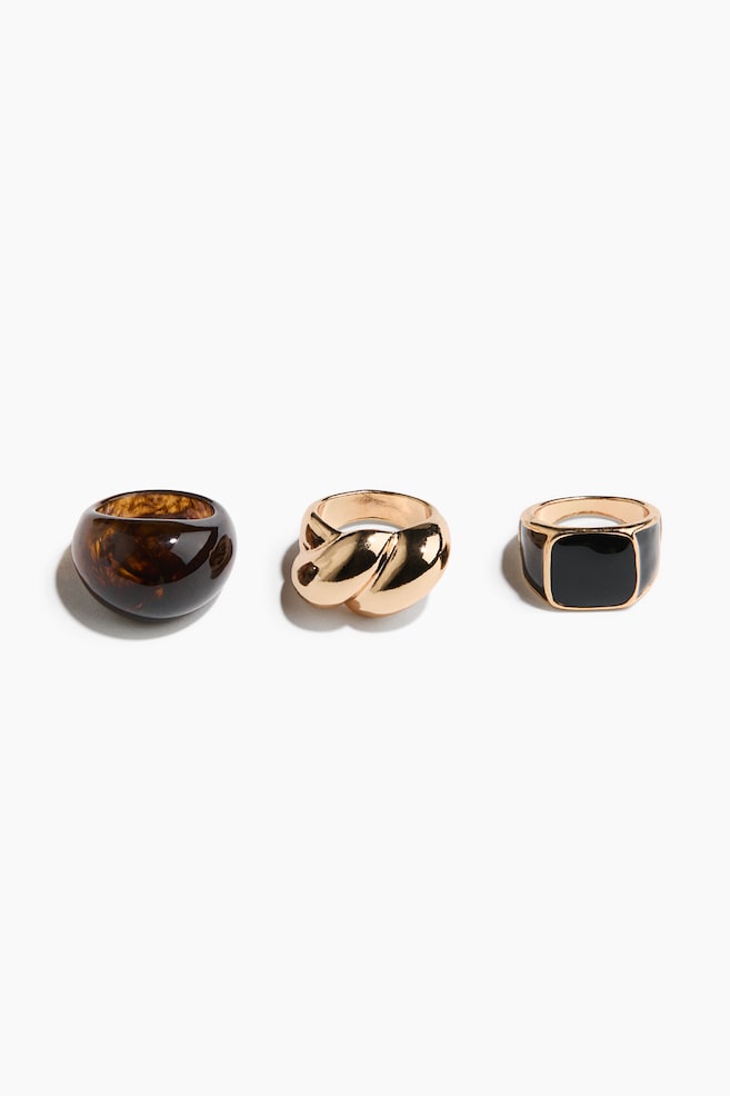 Three chunky rings are featuring various designs, all facing slightly to the right. A rounded tortoiseshell resin ring is resting on the left. A twisted gold-toned metal ring is centered, and a gold-toned metal signet ring with a rectangular black enamel top is positioned on the right. - 1