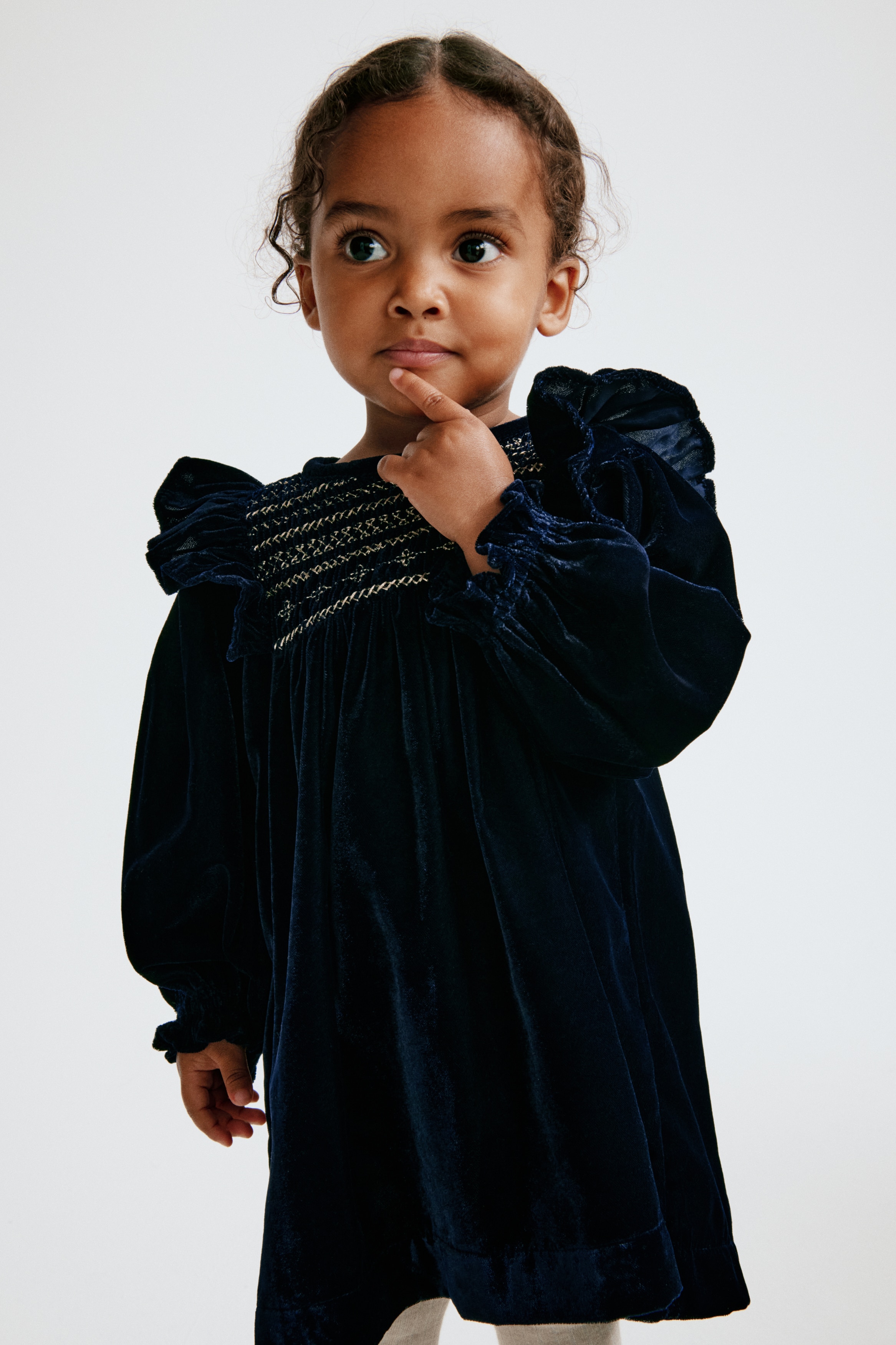 A navy blue velvet baby dress is facing front, showcasing a smocked chest panel with gold embroidered patterns and ruffled trim at the shoulders. The dress features long sleeves with elasticised cuffs and a gathered skirt that flows from the bodice.