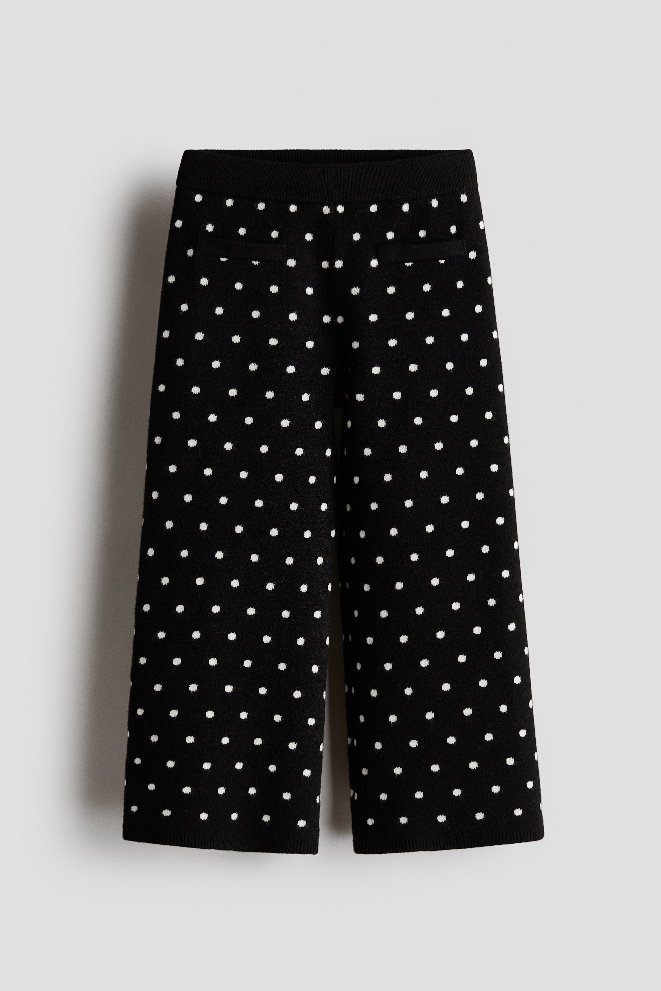 Agrandir l'image: Une paire de pantalons noirs en maille, cropped et à jambes larges, est suspendue de face, avec un motif à pois blancs sur l’ensemble. Le pantalon est doté d’une taille côtelée et de deux poches passepoilées horizontales près du haut.