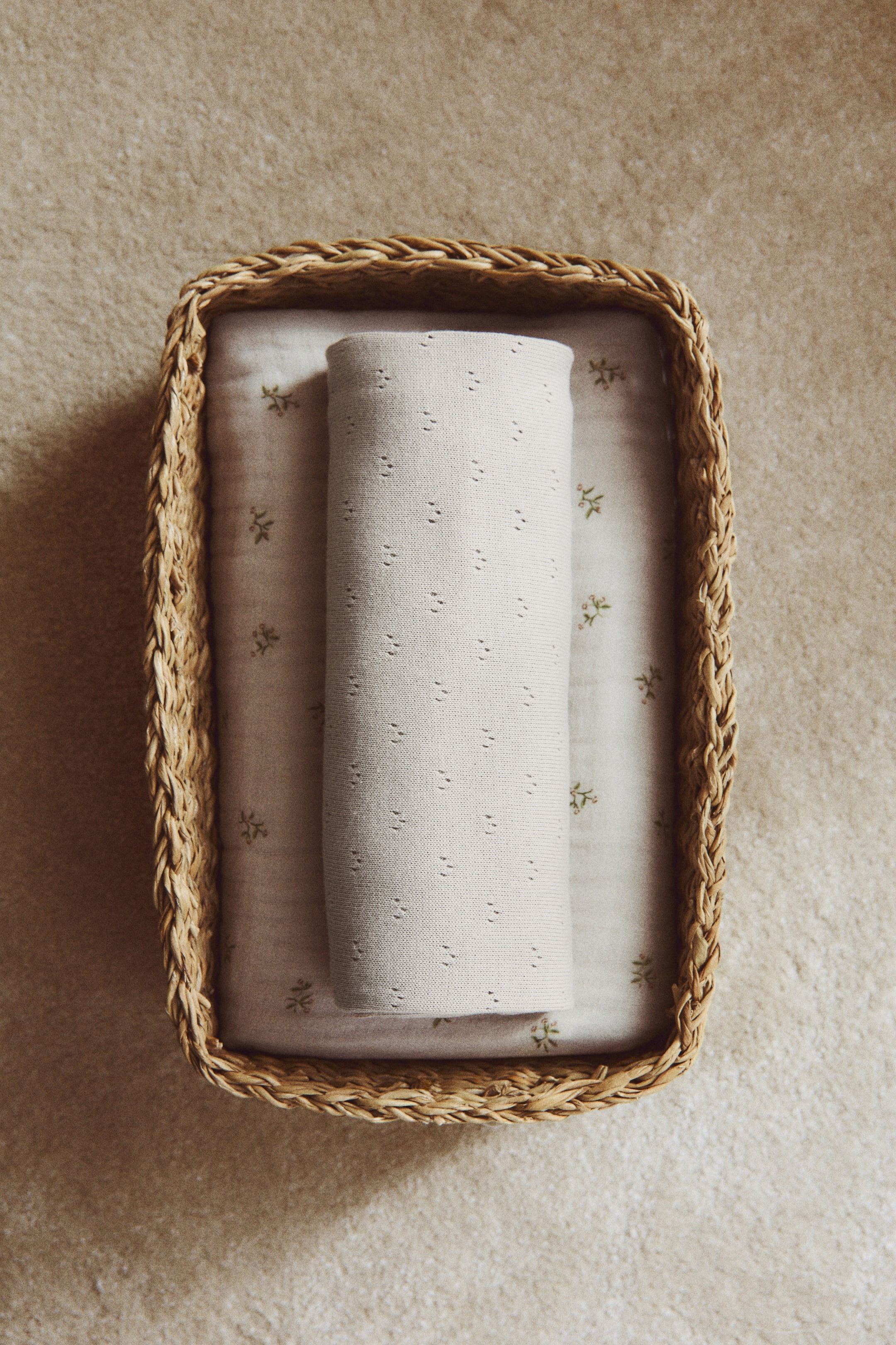 Voir une image plus grande: Une couverture pour bébé de couleur crème avec un tricot pointelle est roulée dans le sens de la longueur, reposant sur un tissu beige plus clair orné de petits motifs botaniques verts, le tout contenu dans un panier rectangulaire en osier.