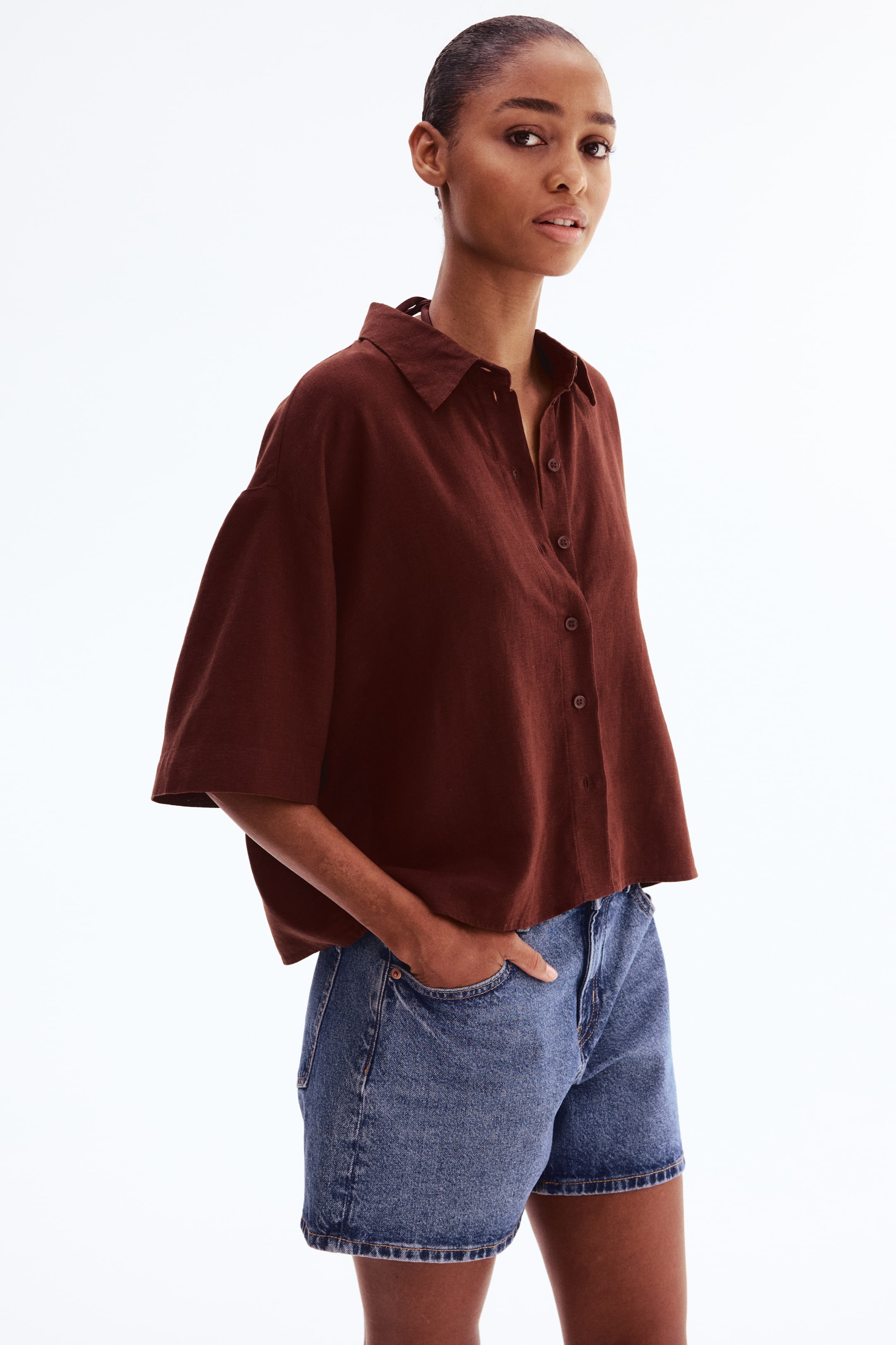 View larger image: A reddish-brown shirt in an airy linen and viscose blend weave is being worn by the model, featuring a collar, a partially unbuttoned front, gently dropped shoulders, and elbow-length sleeves. The model is also wearing blue denim shorts.