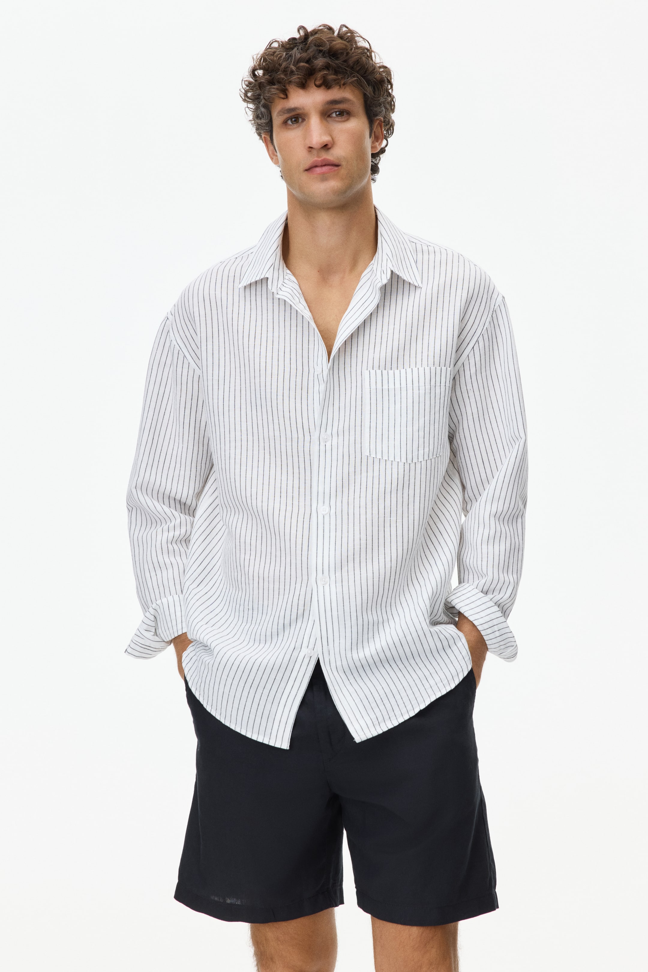 View larger image: A white long-sleeved shirt, featuring thin dark vertical stripes, is worn open over dark shorts, highlighting its relaxed fit, a left chest patch pocket, a flat spread collar, and rolled-up cuffs.