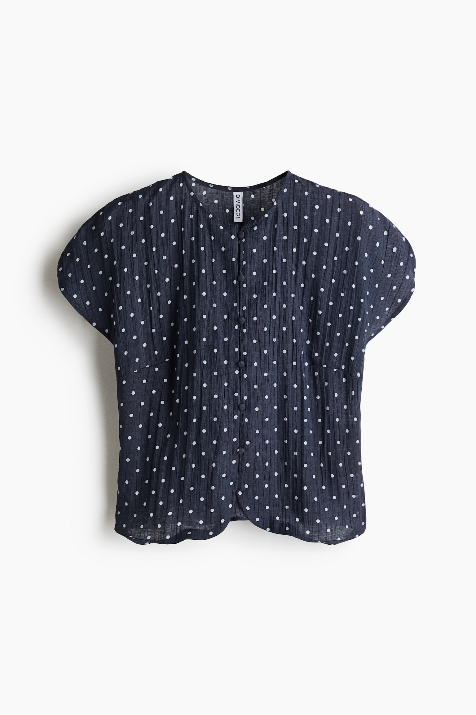A dark navy blue short-sleeved blouse is shown from the front, featuring a white polka-dot pattern on a crinkled-texture fabric. The top is designed with a collarless round neckline, a central button-through closure with matching fabric-covered buttons, and short, wide cap sleeves.
