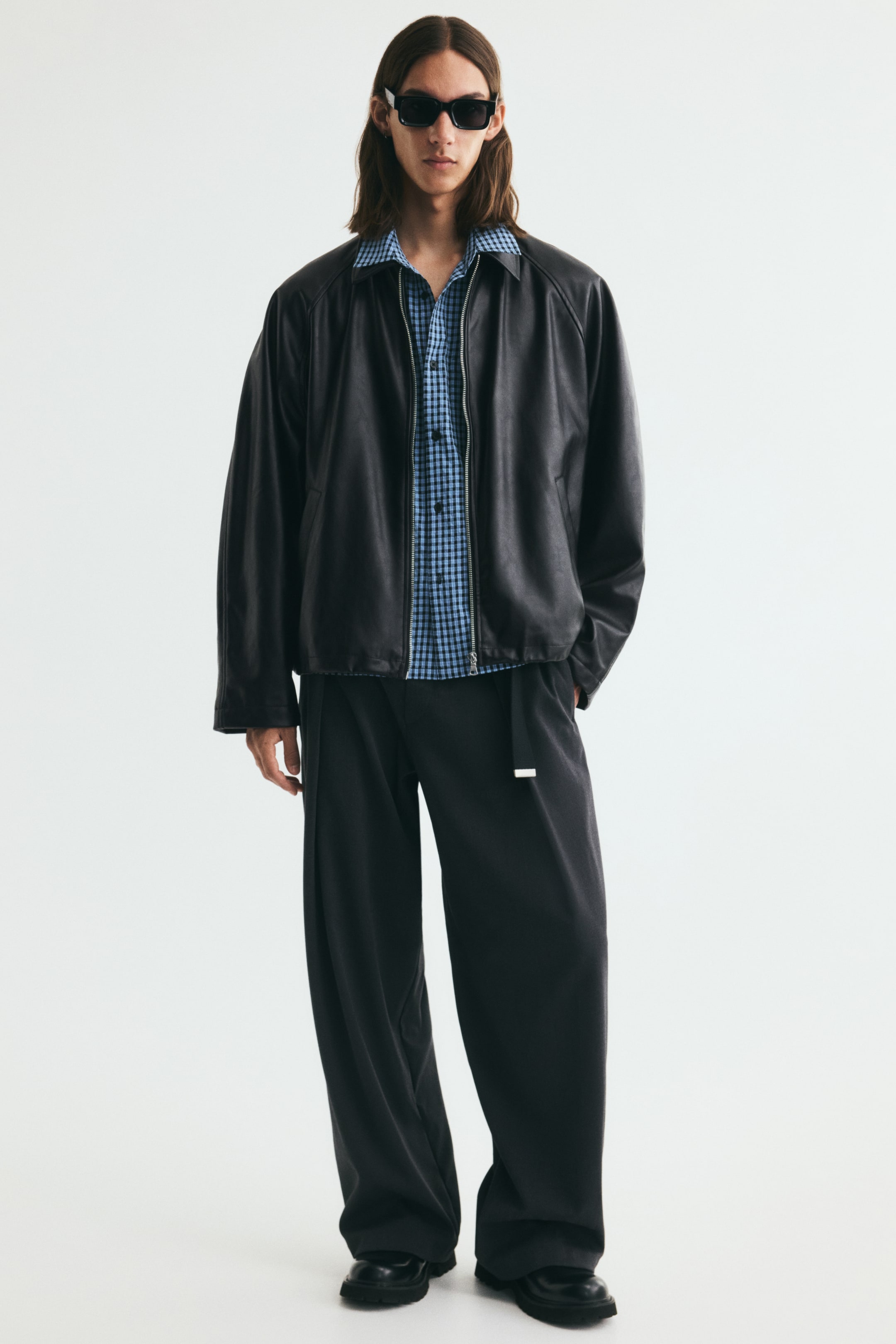 View larger image: A black jacket in coated fabric featuring a collar, a front zipper, and raglan sleeves is worn open by the model. The jacket is layered over a blue and black checkered shirt and styled with loose-fitting black trousers.