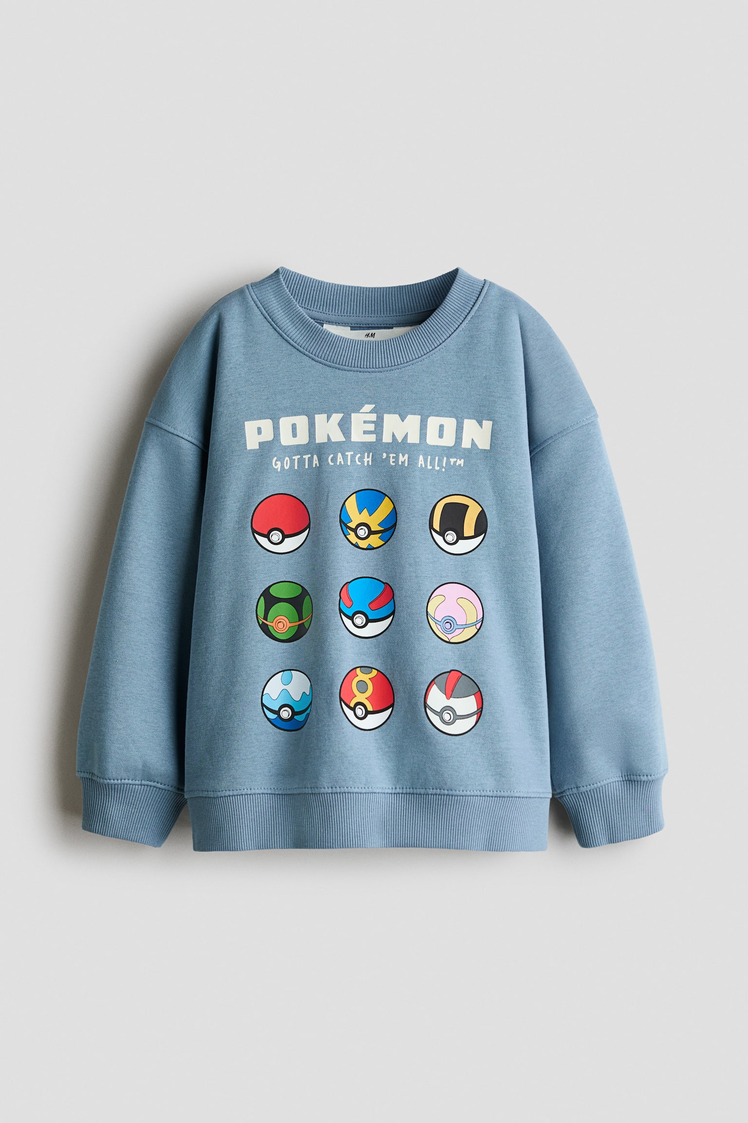 Un sweat-shirt bleu poussiÃ©reux est prÃ©sentÃ© de face, mettant en valeur un col rond cÃ´telÃ©, des Ã©paules tombantes, ainsi que des poignets et un ourlet cÃ´telÃ©s. La poitrine affiche un texte graphique blanc indiquant Â« POKï¿½MON Â» et Â« GOTTA CATCH 'EM ALL!? Â» au-dessus d'une grille de neuf Pokï¿½ Balls multicolores.